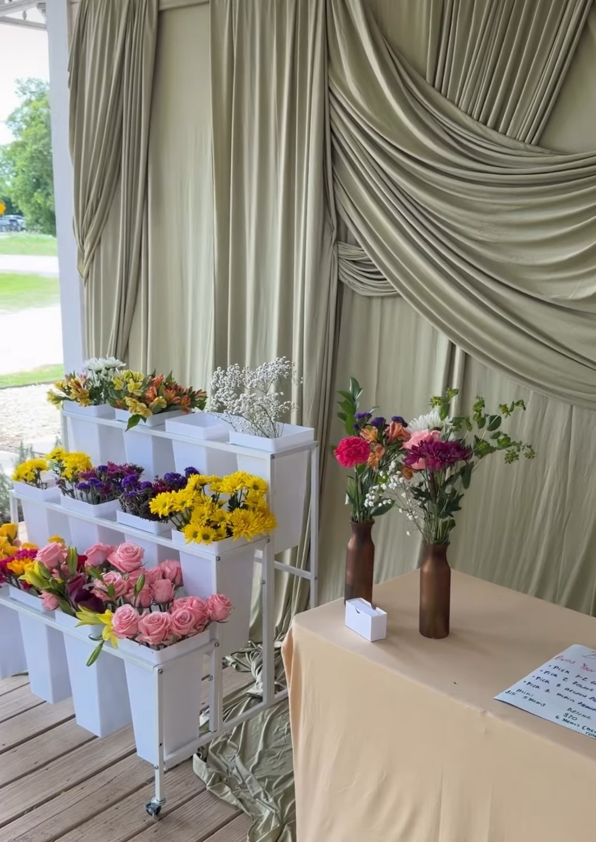 Curtains with draped fabric, flower arrangements in vases, and a table with a beige cloth and a paper, set outdoors.