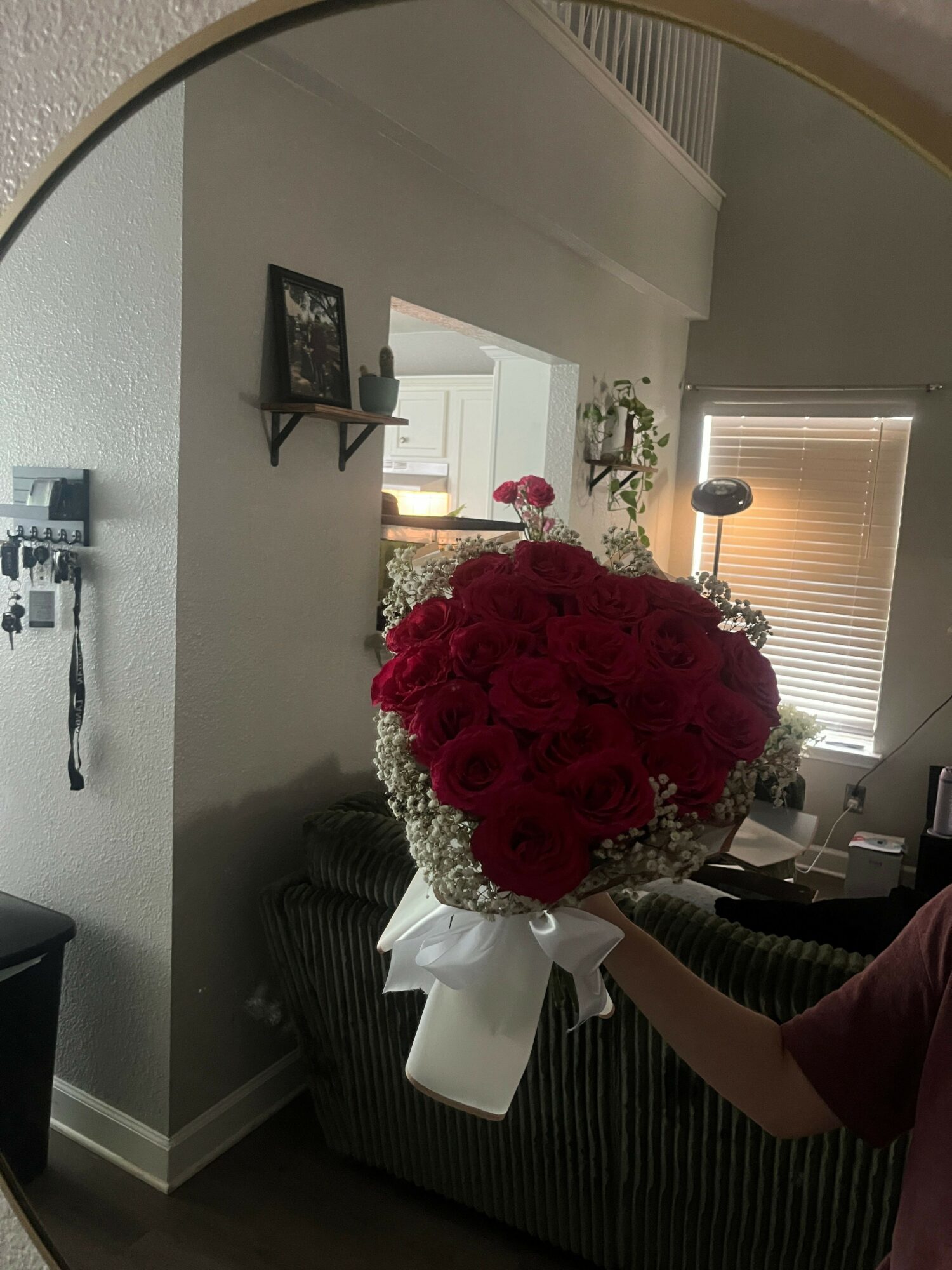 A large bouquet of red flowers in a white vase on a table, with a person partially visible nearby, in a room with a window and framed photo.