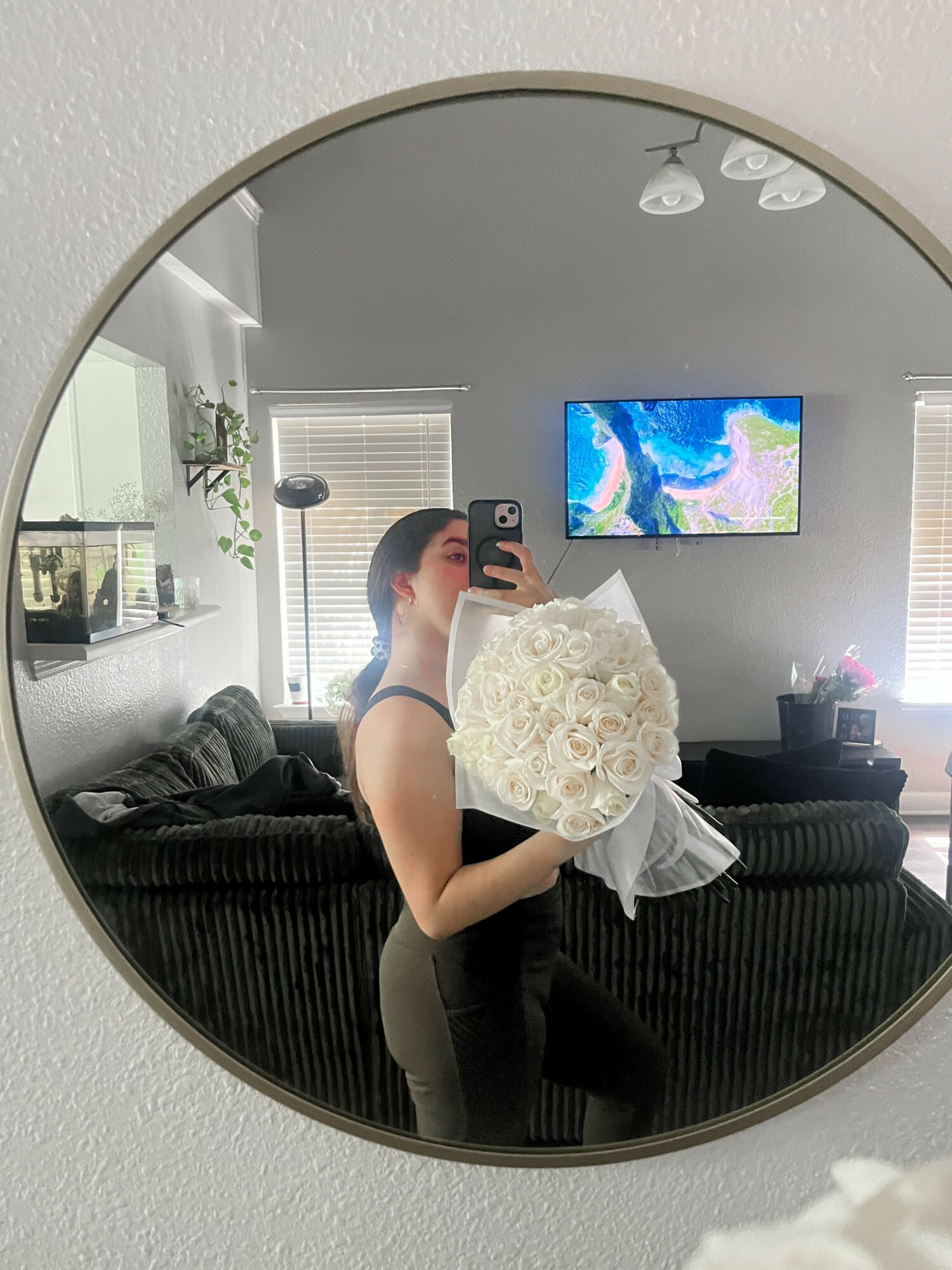 Person holding a bouquet of white roses taking a selfie in a round mirror, living room background, TV on wall.
