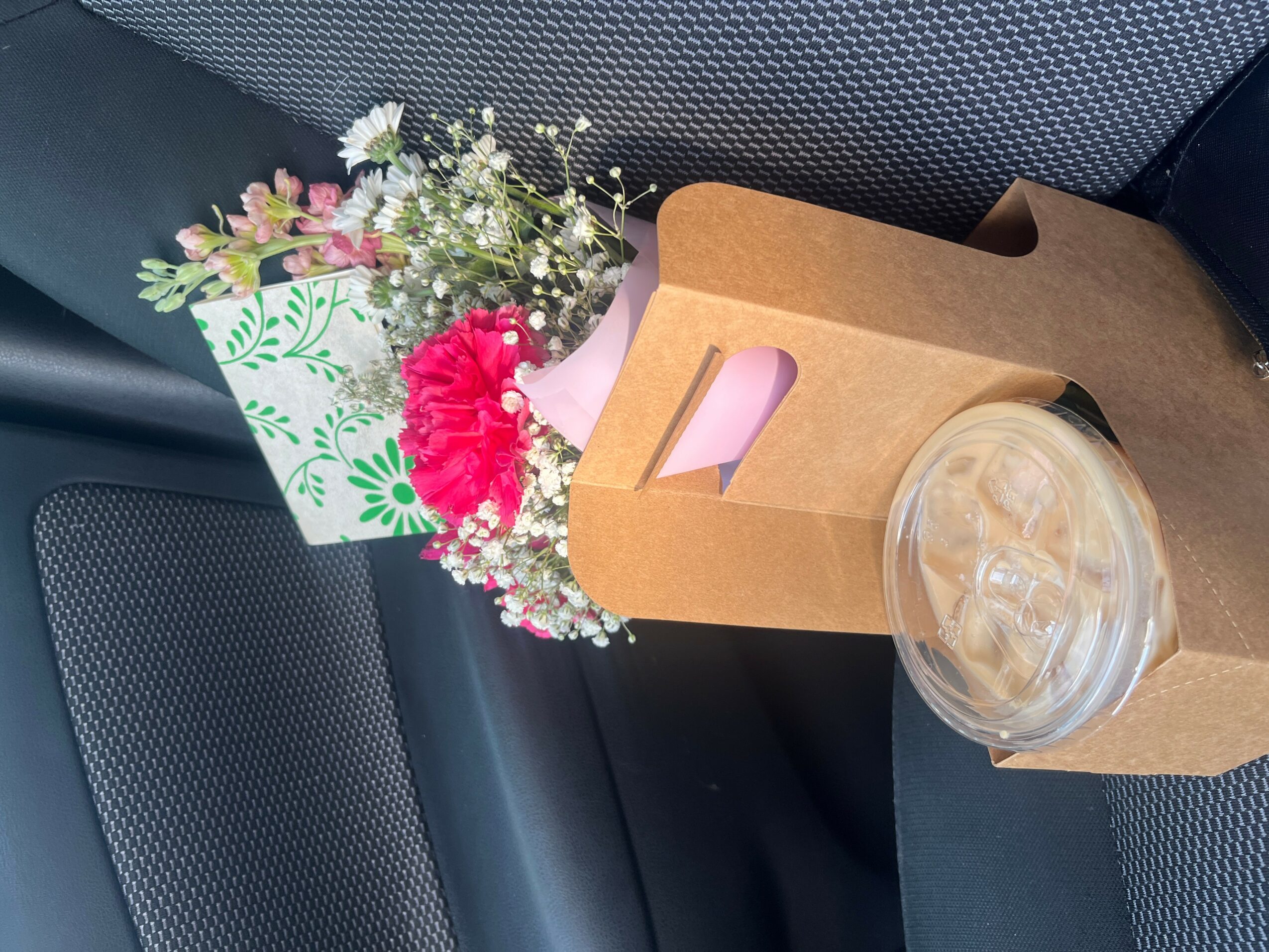 Flowers in a paper bag and a plastic cup with a lid on a car seat.