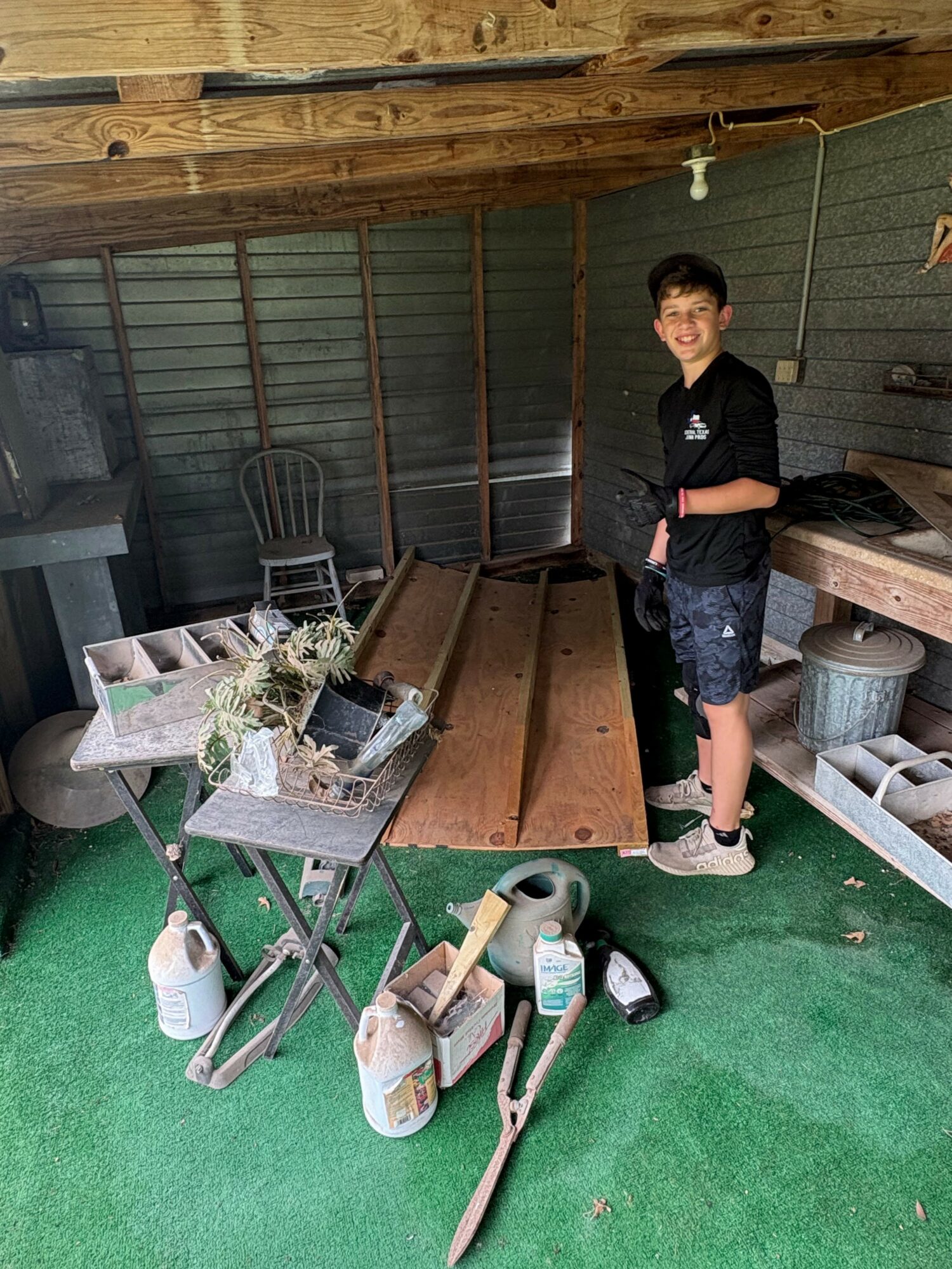 Young boy with dark hair and glasses smiling, standing in a shed with wooden and metal objects, green floor, and tools.