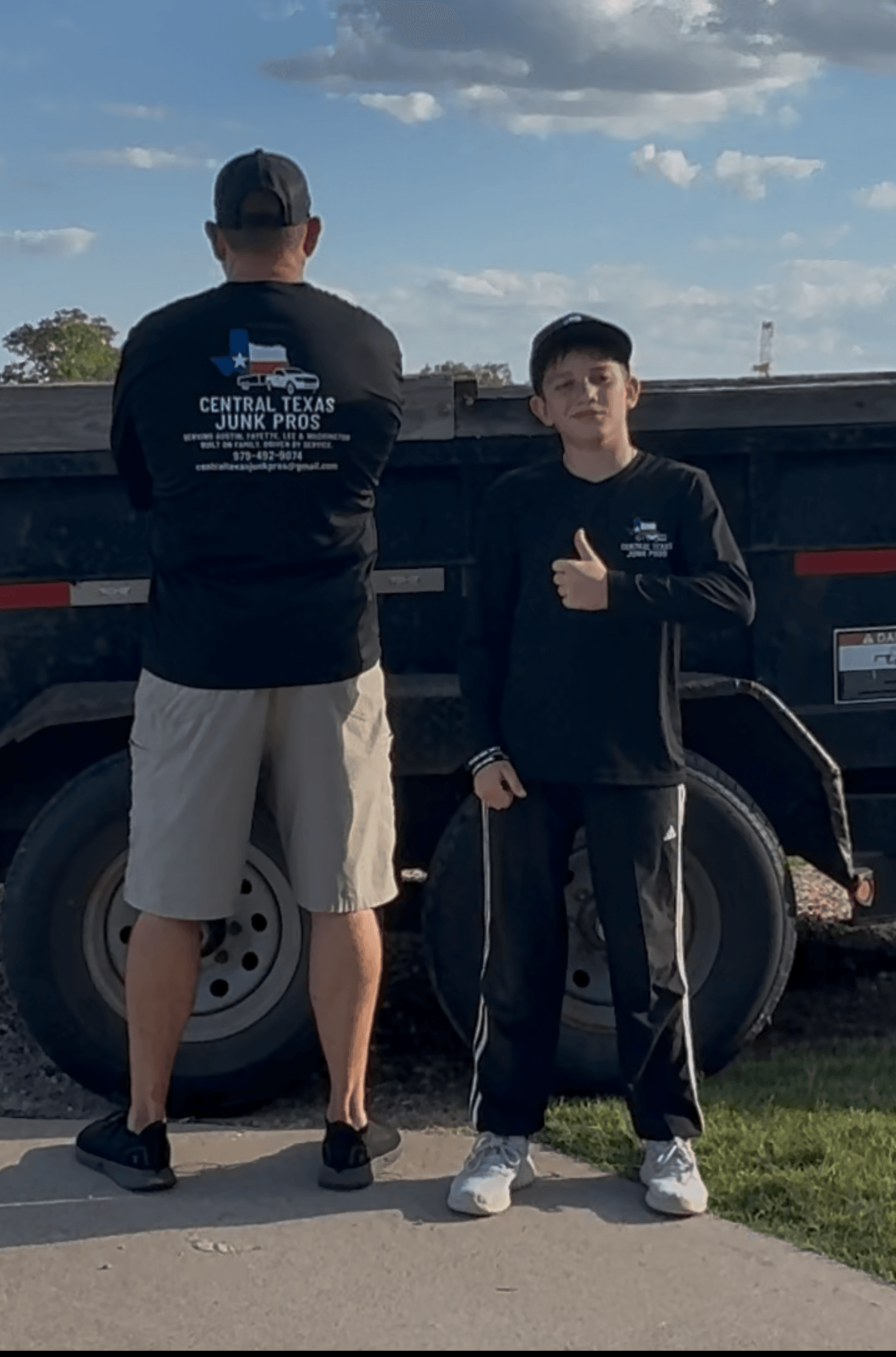 Two people standing outdoors near a large vehicle, one facing away and the other facing forward, under a partly cloudy sky.