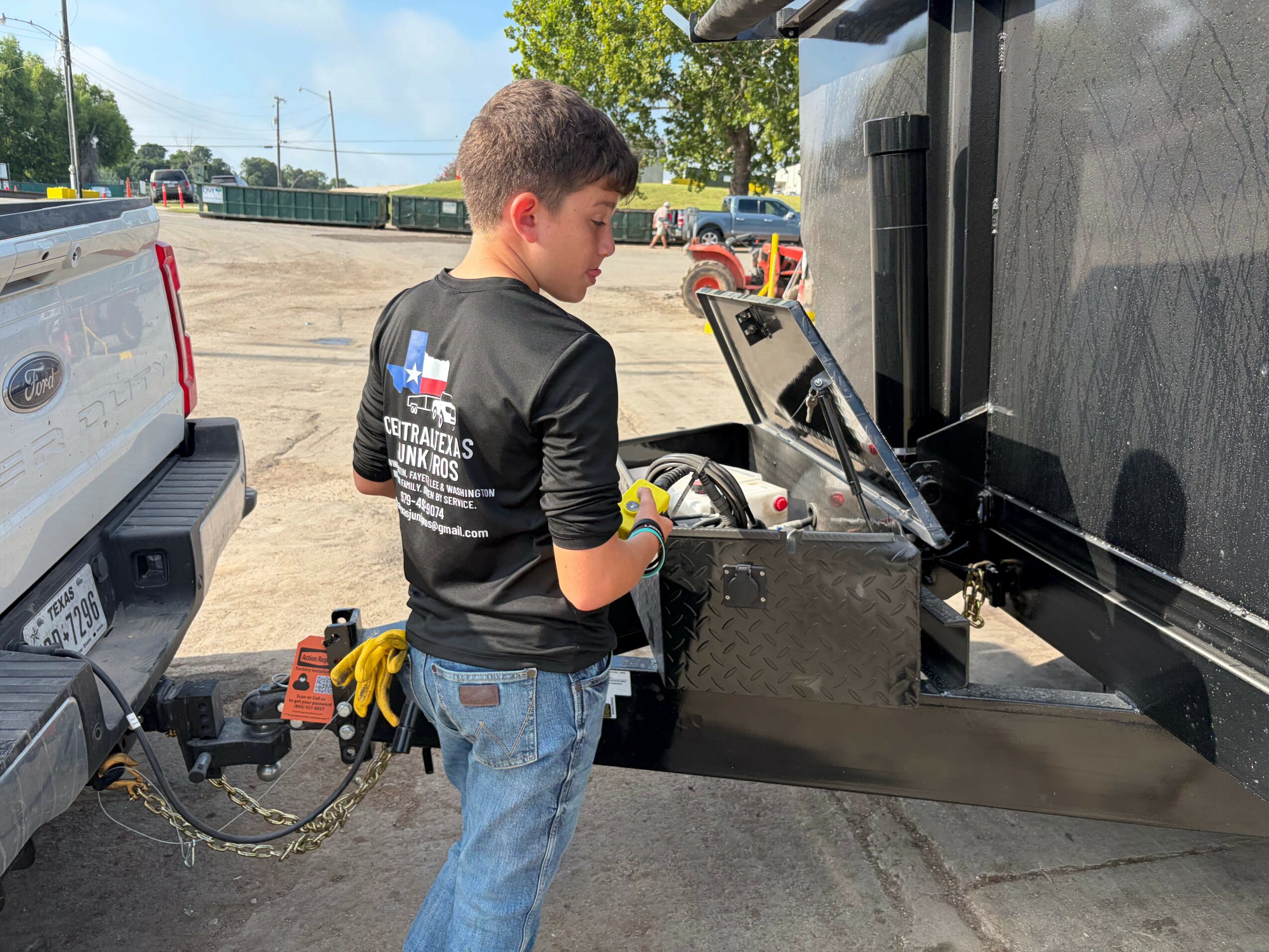 Young boy in black shirt with logo, working with tools near a black equipment box, outdoors in a parking lot.