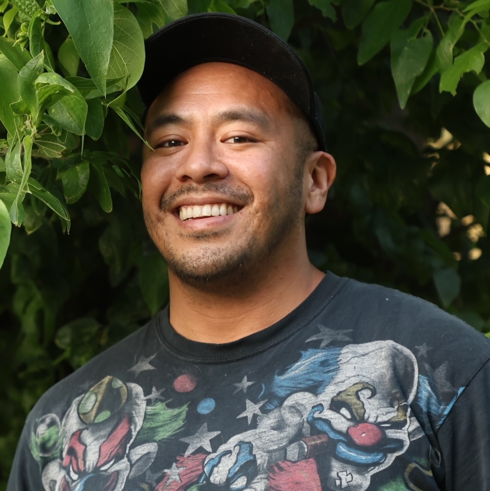 Smiling man wearing a black cap and a graphic t-shirt outdoors with green foliage in the background.