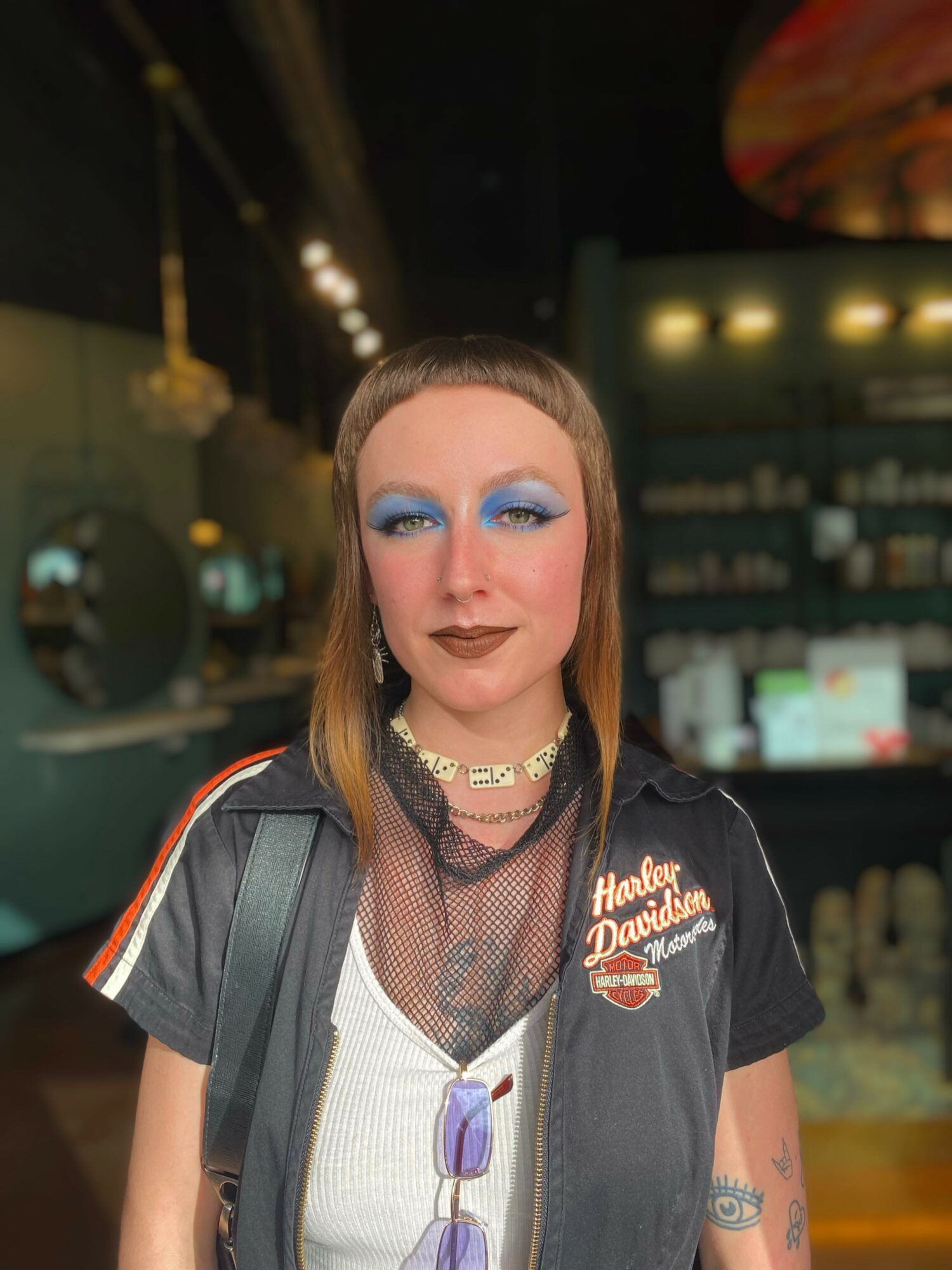 Person with blue eyeshadow, dark lipstick, wearing a Harley Davidson shirt, in a dimly lit bar or restaurant.