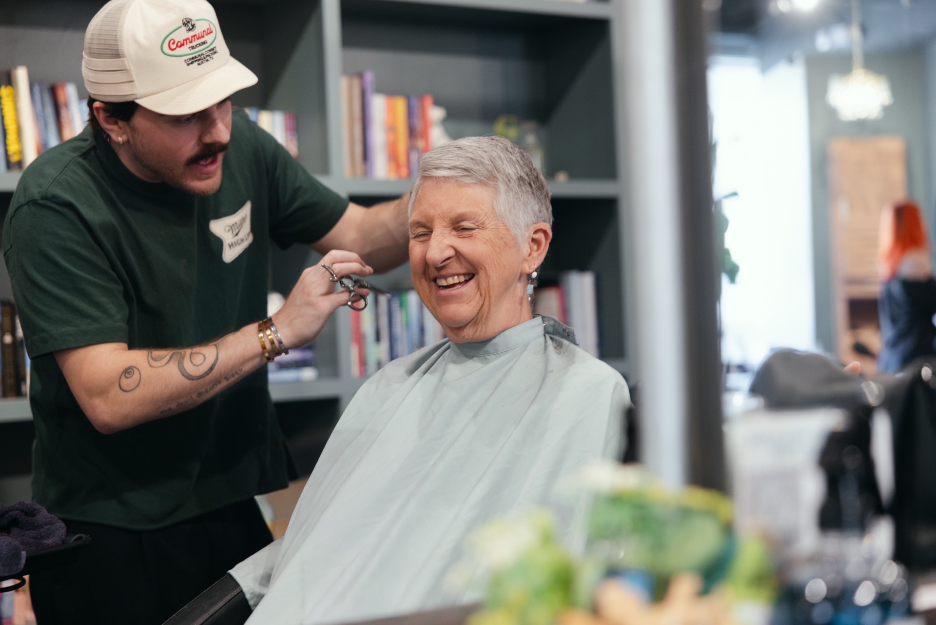 Elderly woman smiling in a barber chair, getting a haircut from a barber in a salon. Shelves and window in background.