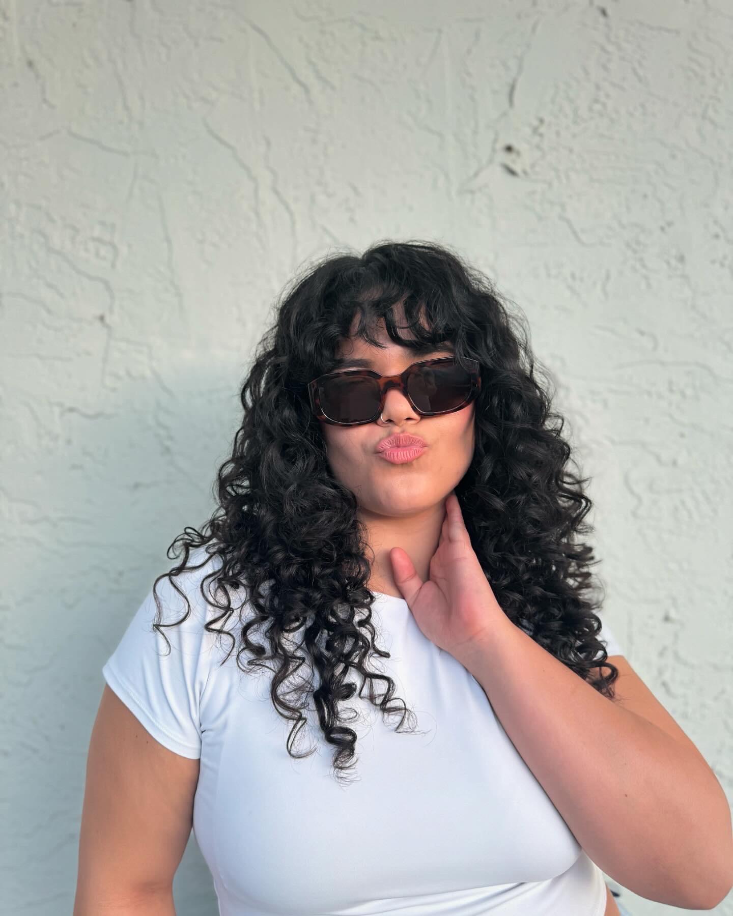 Woman with curly hair wearing sunglasses and a white shirt, posing with hand on neck, against a textured wall.
