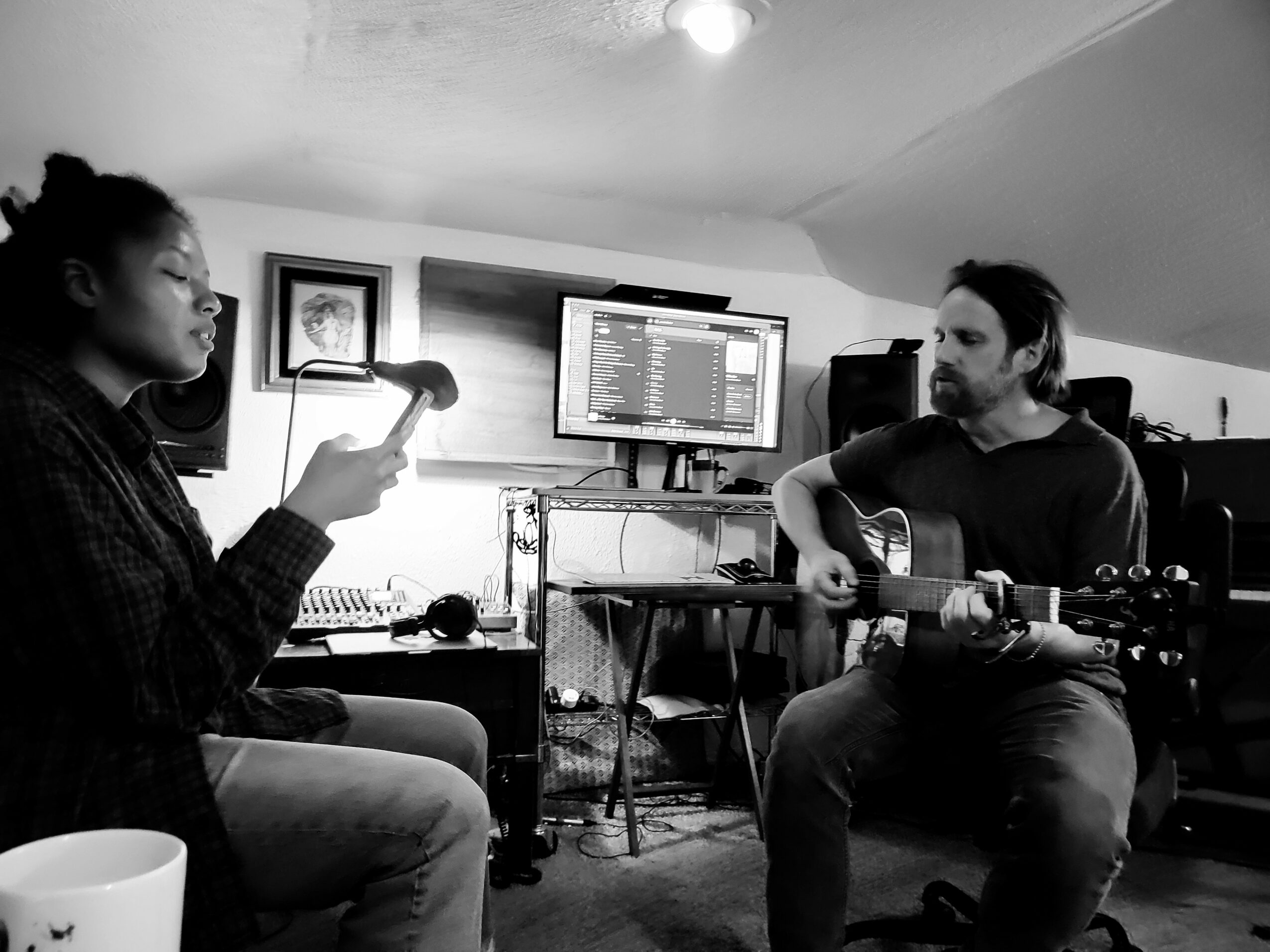 Two people in a music studio, one singing into a microphone and the other playing guitar, with a monitor and equipment in the background.