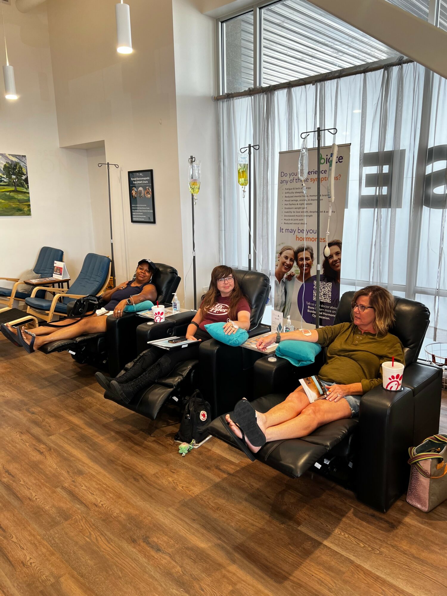 Four people sitting in chairs and recliners in a waiting area, some using phones, with large windows and curtains behind them.