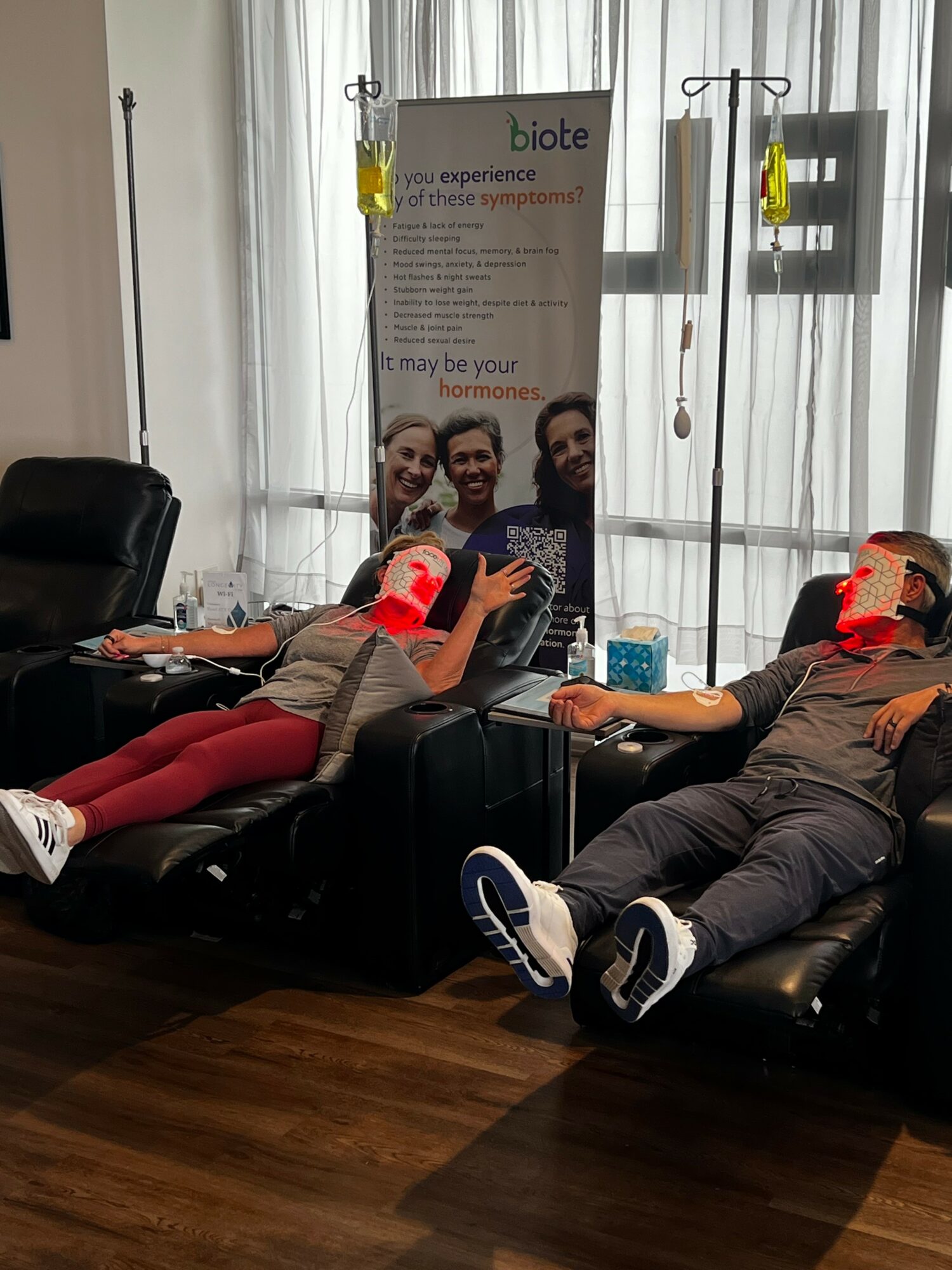 Two people lying on black recliners donating blood, with a banner and window behind them, in a room with wooden floor.