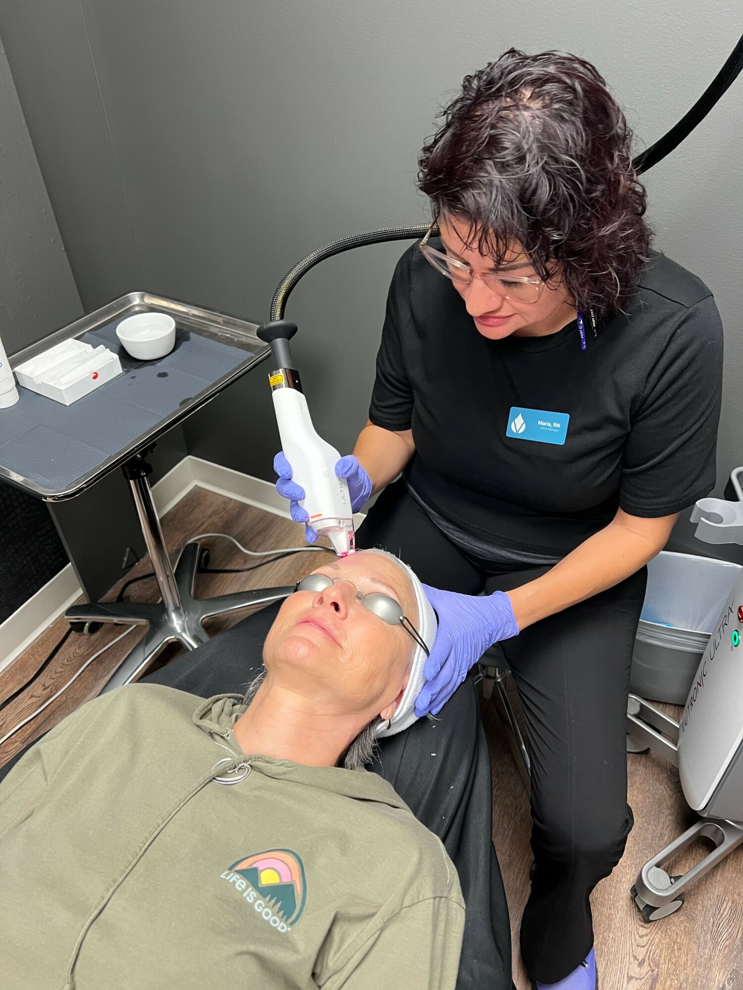 A woman with curly hair performs a procedure on a person lying down, using a handheld device, in a clinical setting.