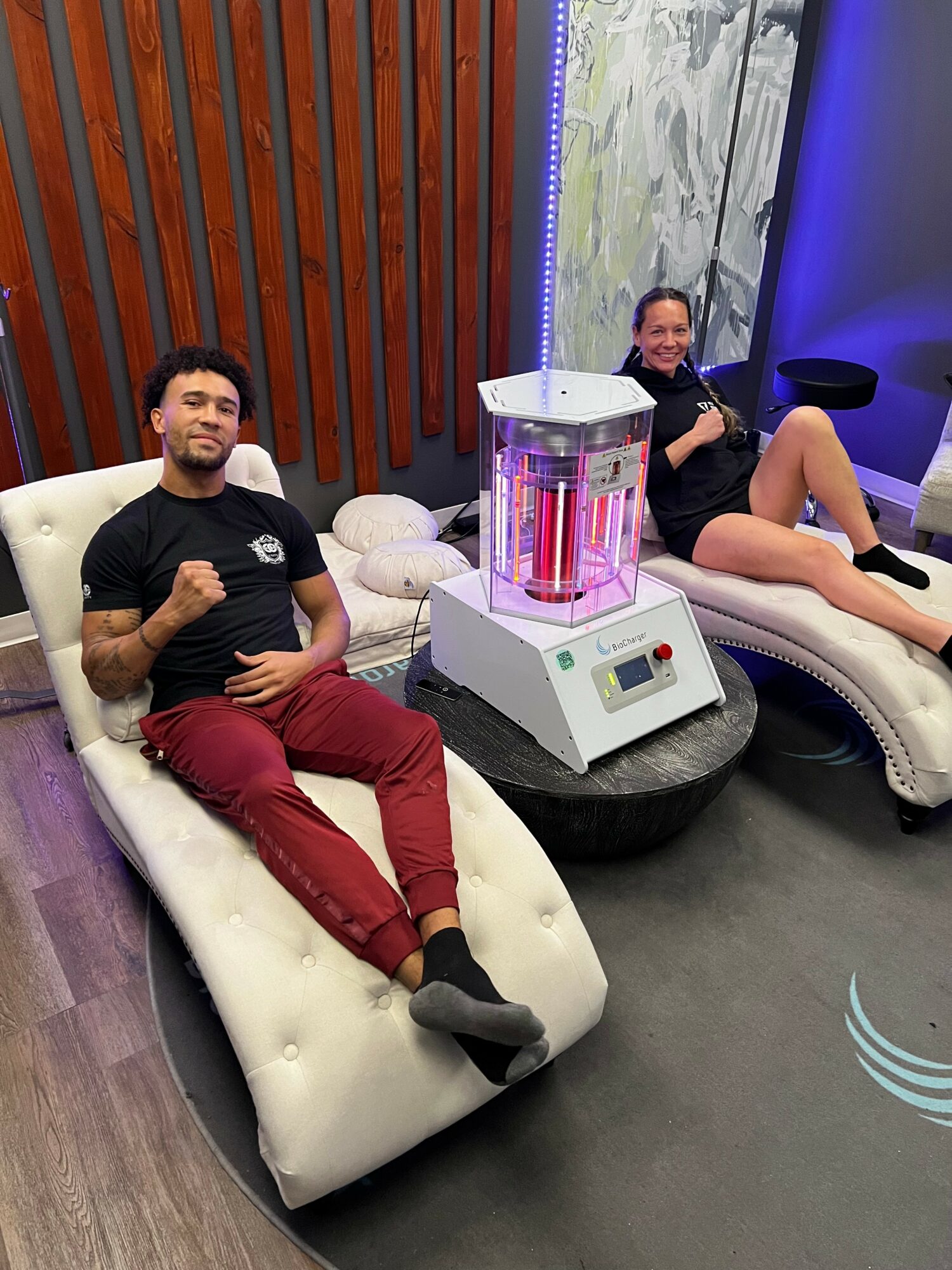 Two people relaxing on lounge chairs with a machine between them, in a room with wooden and marble wall decor.