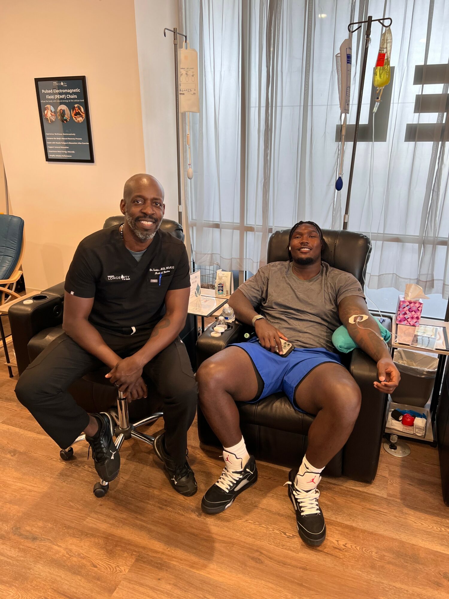 Two men in a medical setting, one sitting in a chair and the other in a recliner, smiling. Medical equipment and curtains in background.