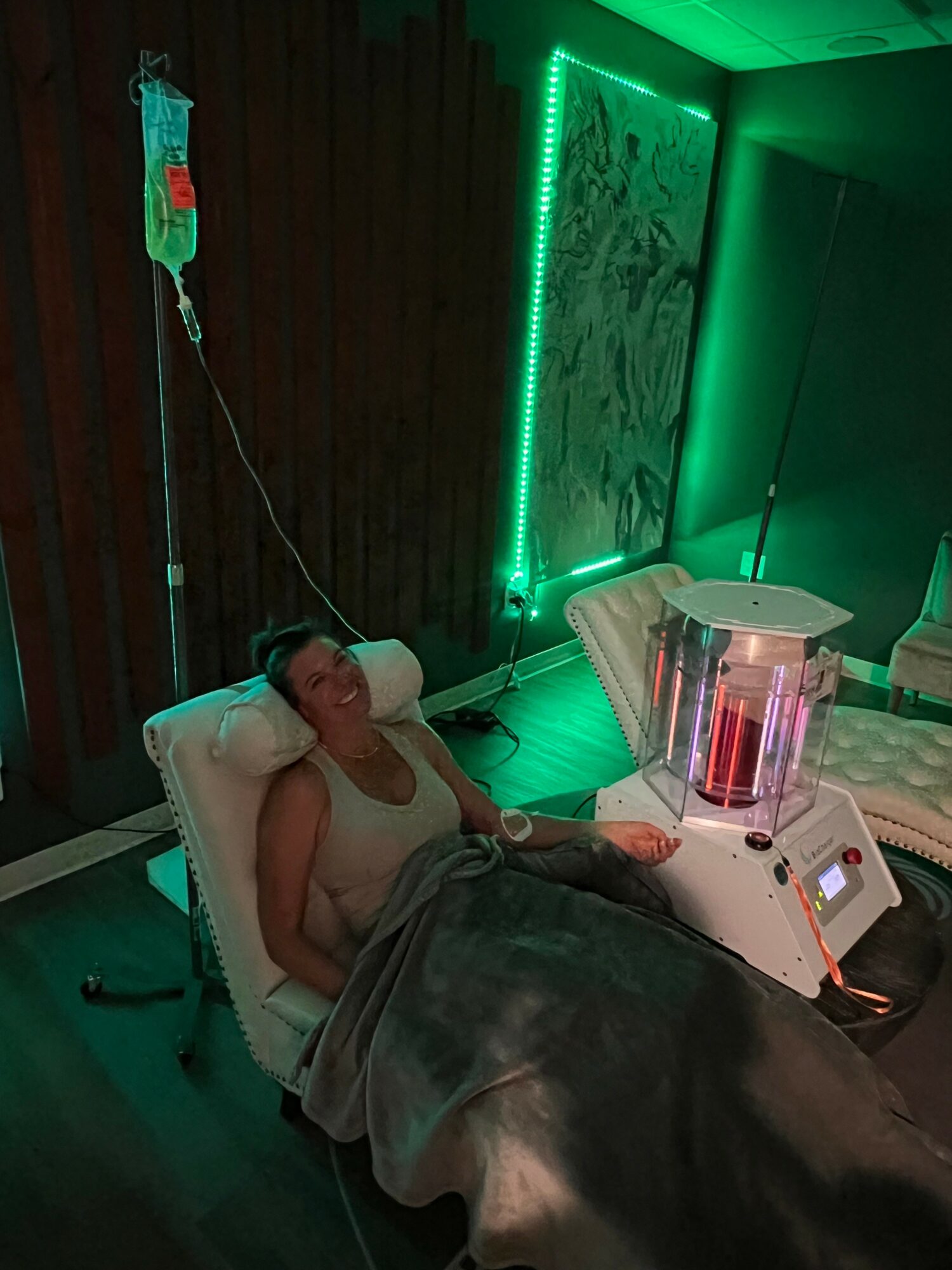 Woman lying in a medical or spa chair connected to a machine with tubes, in a room with green lighting and wall art.