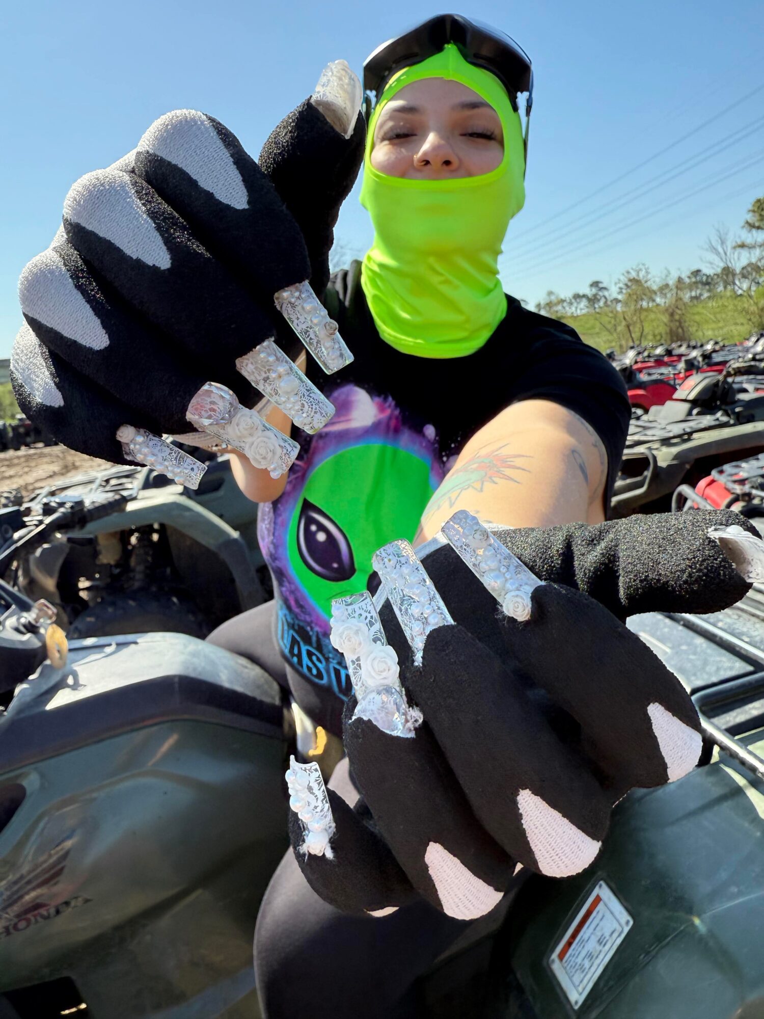 Person wearing a bright green face covering and black gloves, taking a selfie outdoors with a motorcycle and parking lot in background.