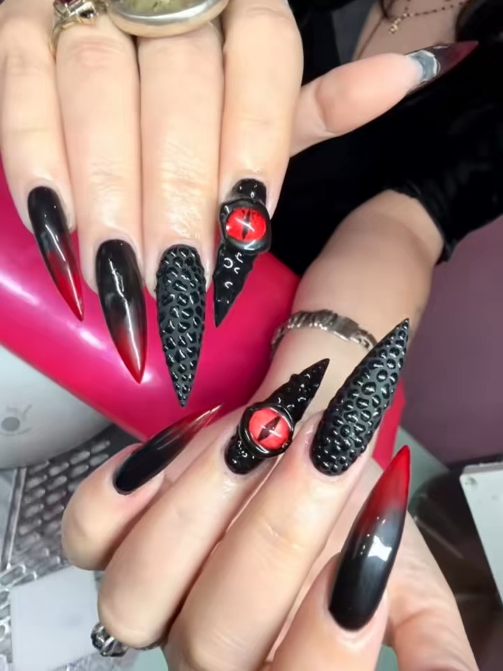 Two hands with long, pointed black and red nails, some with textured or decorative designs, holding a pink object.