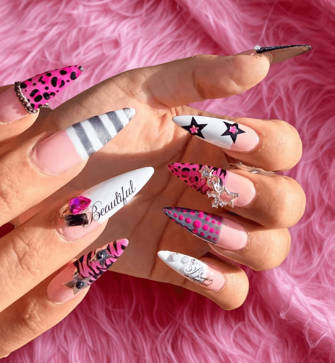 Close-up of hands with colorful, decorated nails against pink furry background, showing various nail art designs.