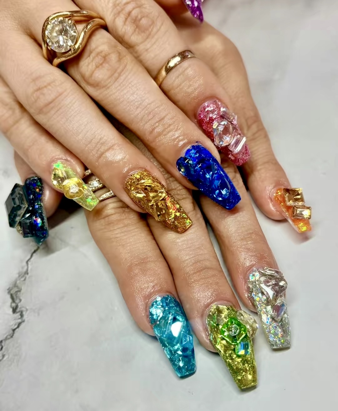 Hands with colorful, decorated nails and multiple rings, resting on a white surface.