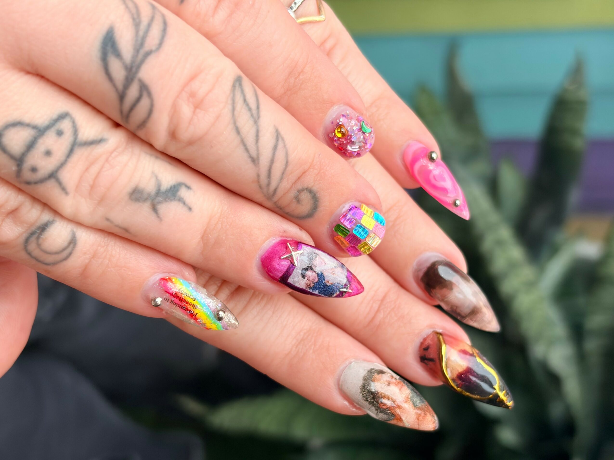 Close-up of a hand with decorated nails and tattoos, outdoors with plants in the background.