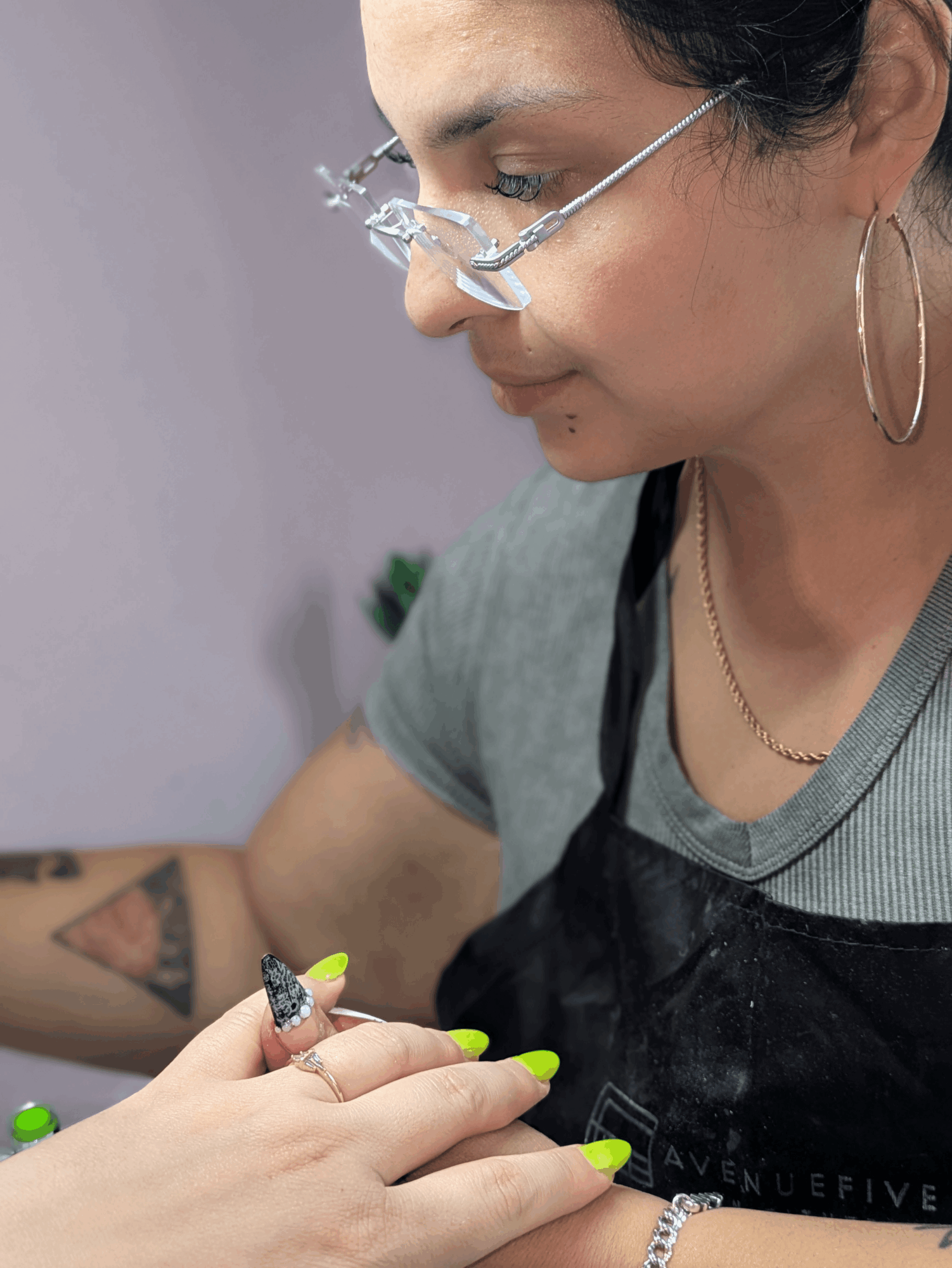 Woman with glasses and hoop earrings, wearing a gray shirt and black apron, holding a small object with green nails.