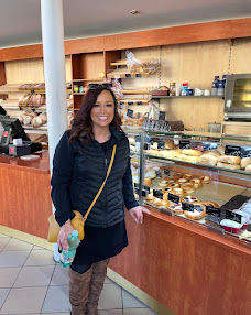 Woman with dark hair smiling in bakery display case with various baked goods, wearing a dark jacket and skirt, holding a yellow bag.