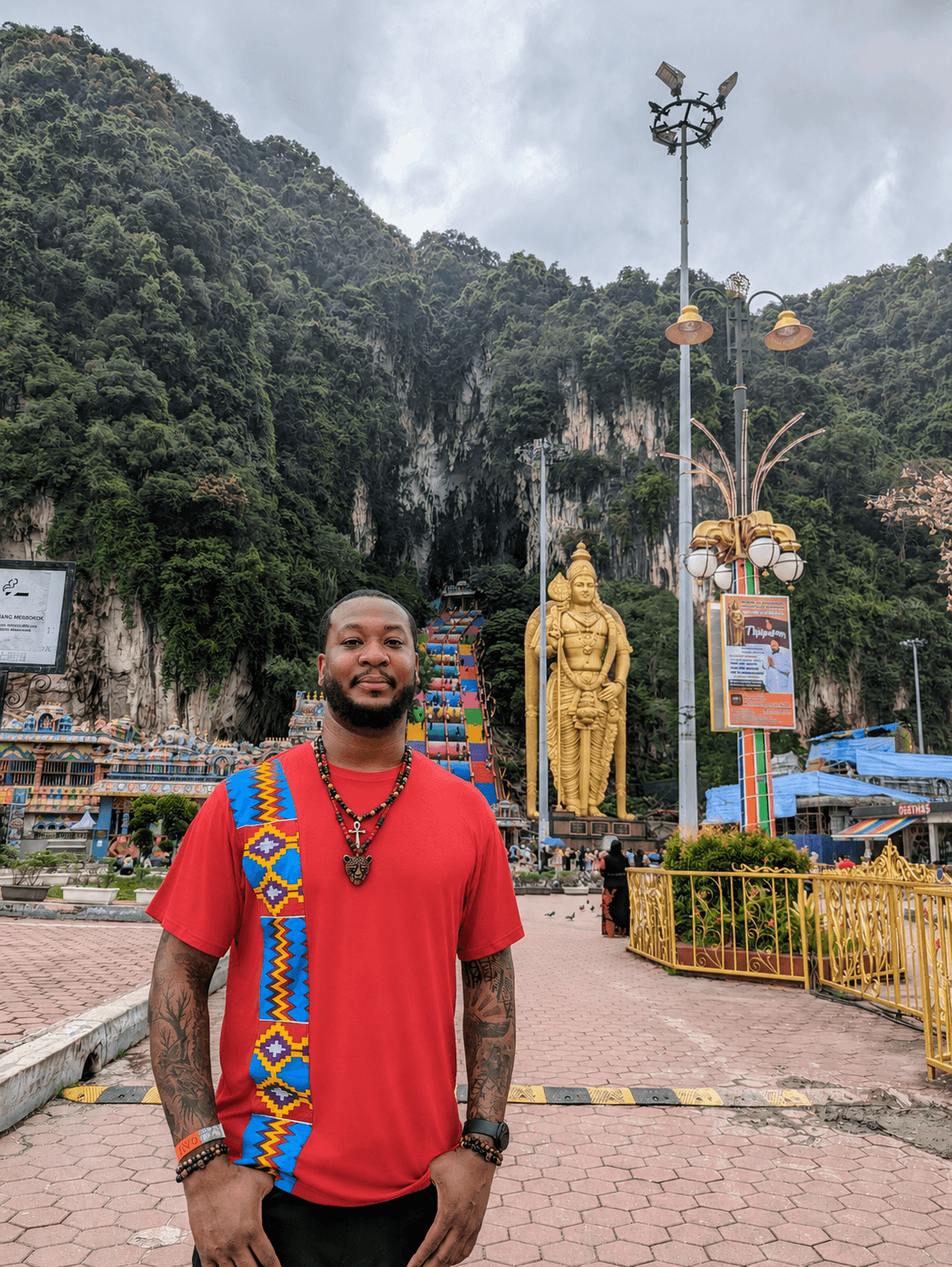 Man in red shirt with colorful sash standing outdoors near a large golden statue and lush green mountains in the background.