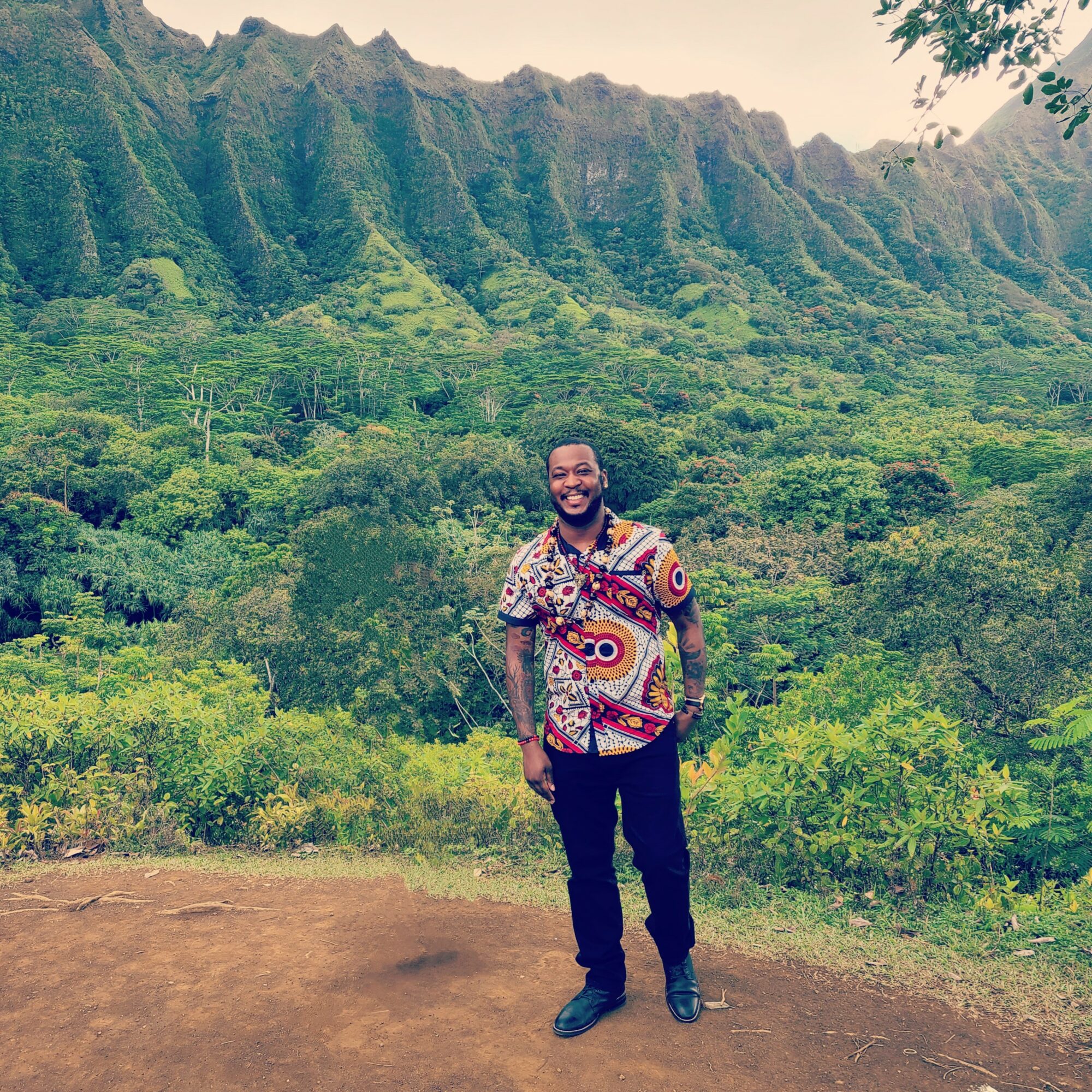 Man standing outdoors in front of lush green mountains, smiling, wearing a colorful patterned shirt and dark pants.