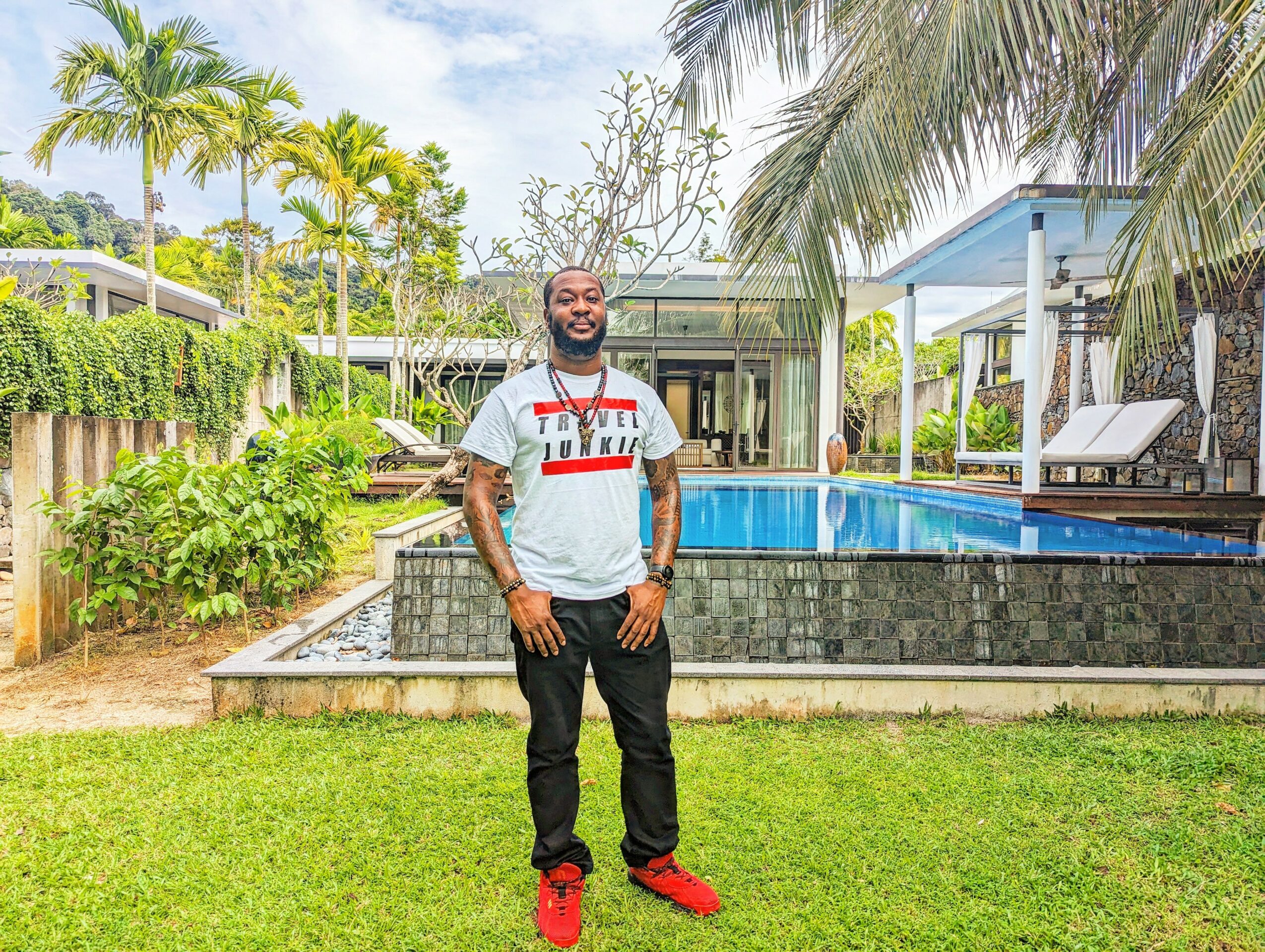 Man standing on grass near a swimming pool with palm trees and modern buildings in background.