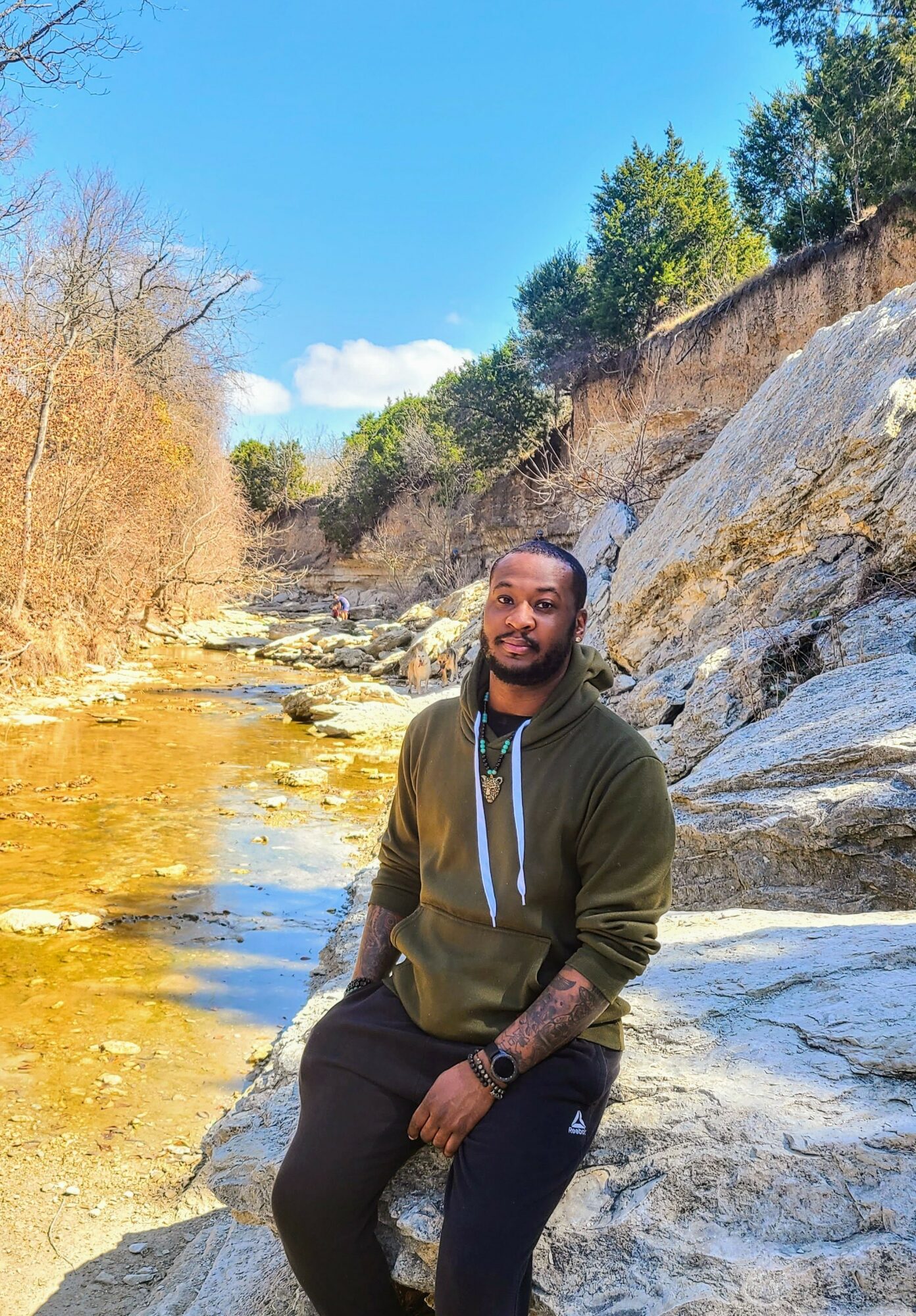 Young man sitting on rocks near a river with a waterfall, trees, and blue sky in the background.