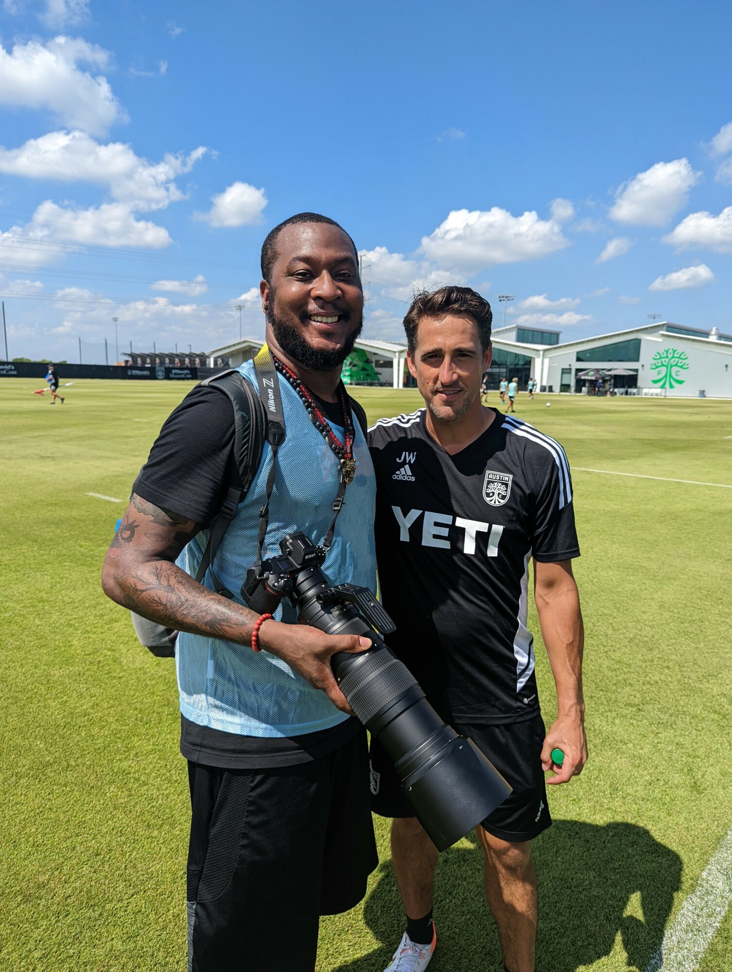 Two men standing outdoors on a grassy field with a blue sky and clouds, one holding a camera with a large lens.