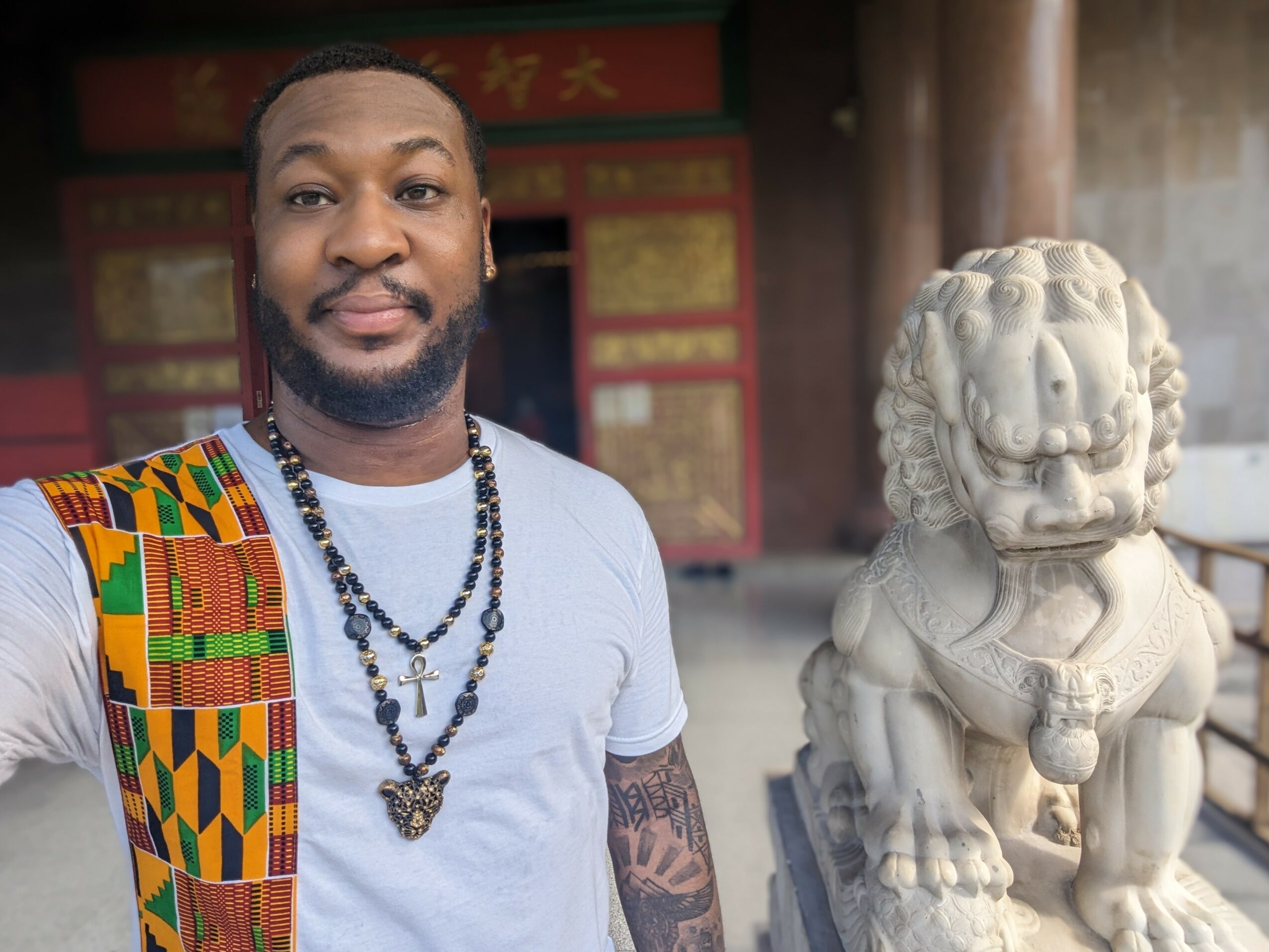 Man taking selfie next to a stone lion statue outside a building with red and gold decorations.