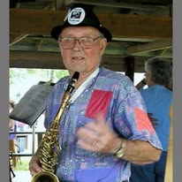 Elderly man wearing glasses, a hat, and a colorful shirt, holding a saxophone, smiling, in a casual outdoor setting.