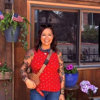 Smiling woman with tattoos wearing a red polka dot top, standing outdoors near a window with potted flowers.