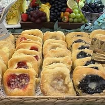 Assorted pastries with fruit fillings arranged in rows, with fruit bowls in the background.