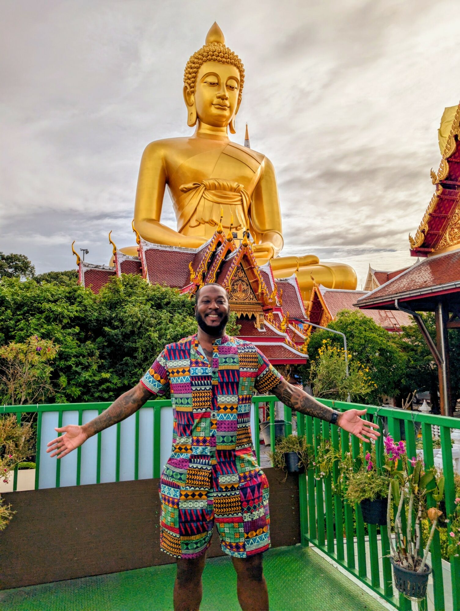Smiling man with arms outstretched standing on a balcony with a large golden Buddha statue in the background.