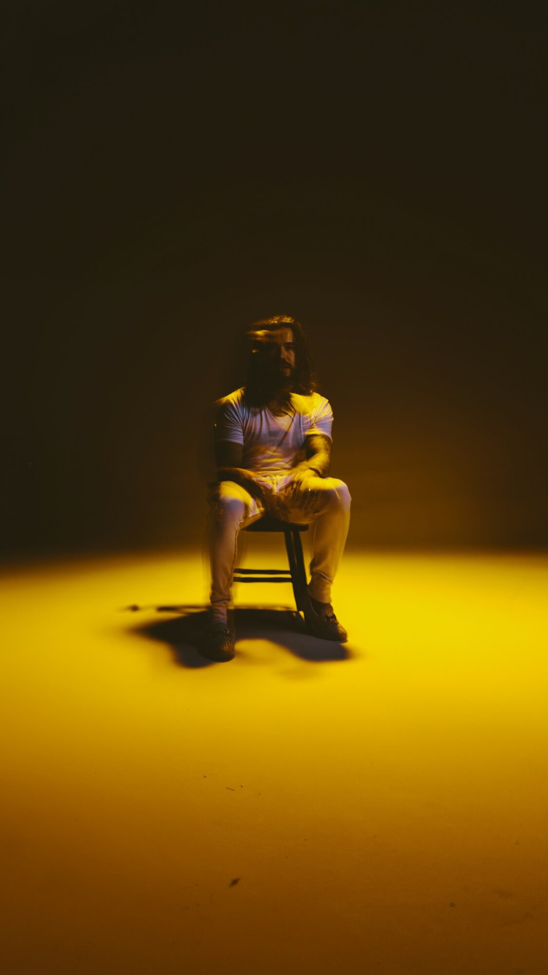 Person sitting on a chair in a spotlight with a dark background, wearing a striped shirt and shorts.