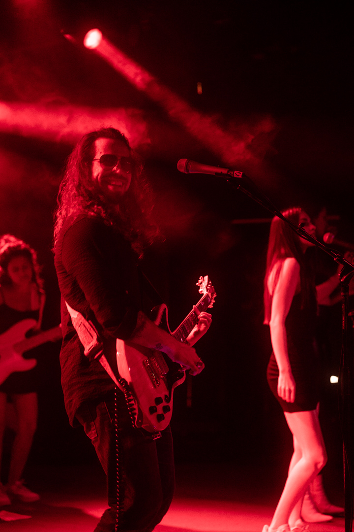 Musicians perform on stage with red lighting, one playing guitar and another singing into a microphone.