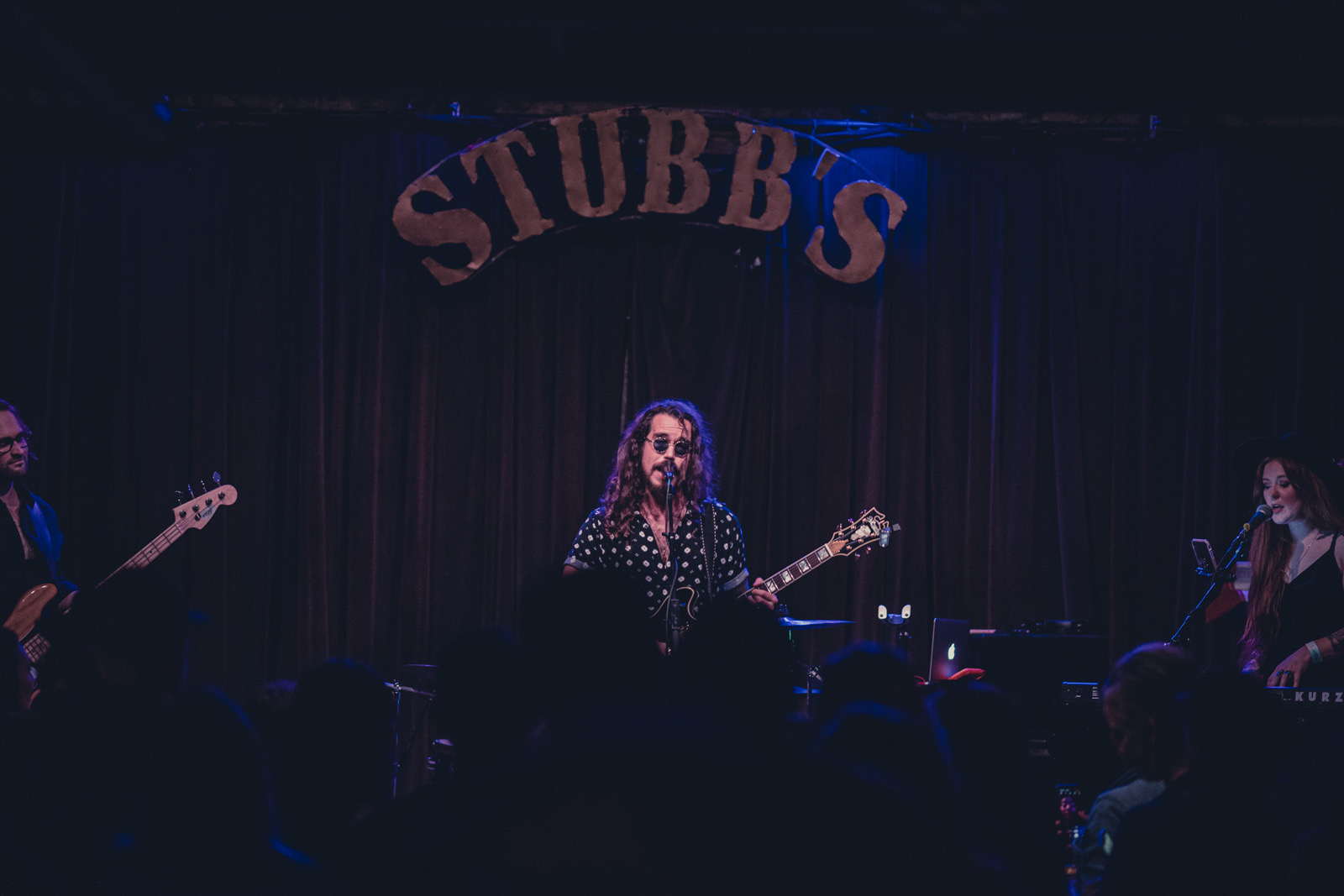 Performer singing and playing guitar on stage with two other musicians, stage backdrop reads 'STUBB'S'.