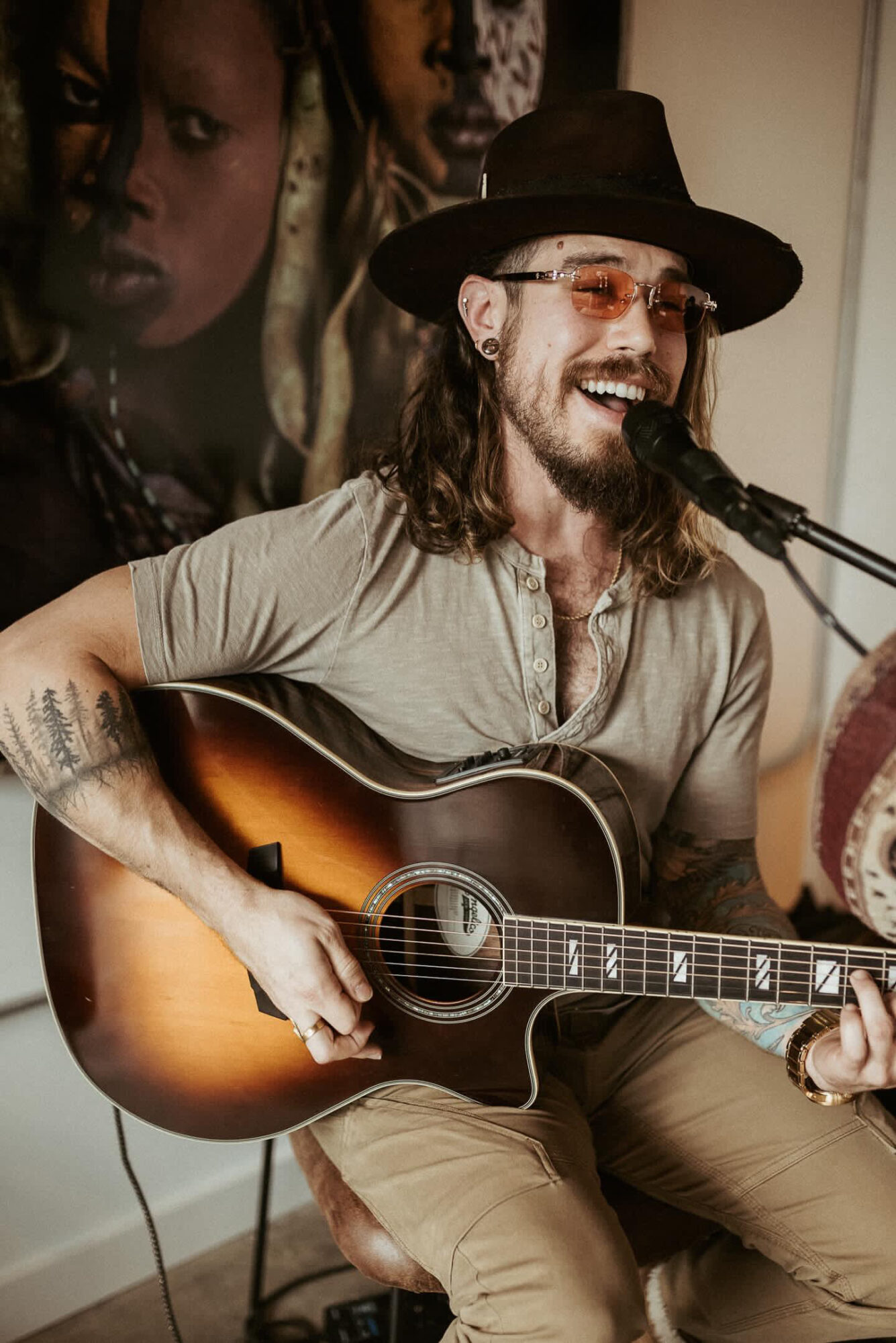 Man with long hair, beard, sunglasses, and hat playing guitar and singing into microphone, smiling.