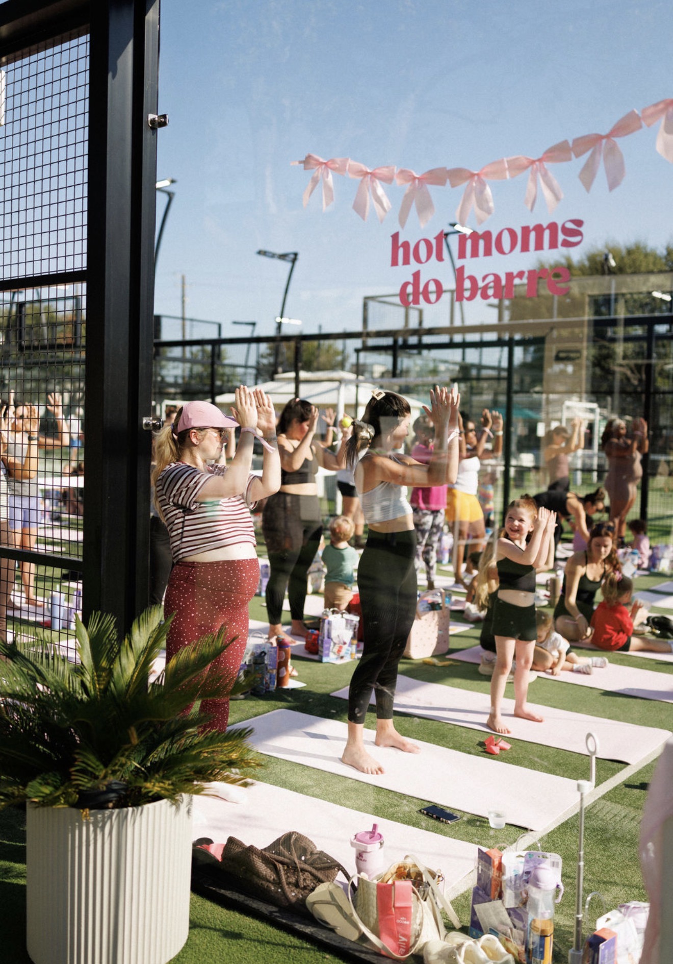 Group of children and adults participating in outdoor yoga class on mats, with pink banners and a sign in the background.