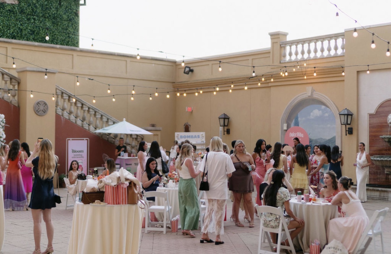 People gather at outdoor event with tables, string lights, and a decorative wall, enjoying food and conversation.