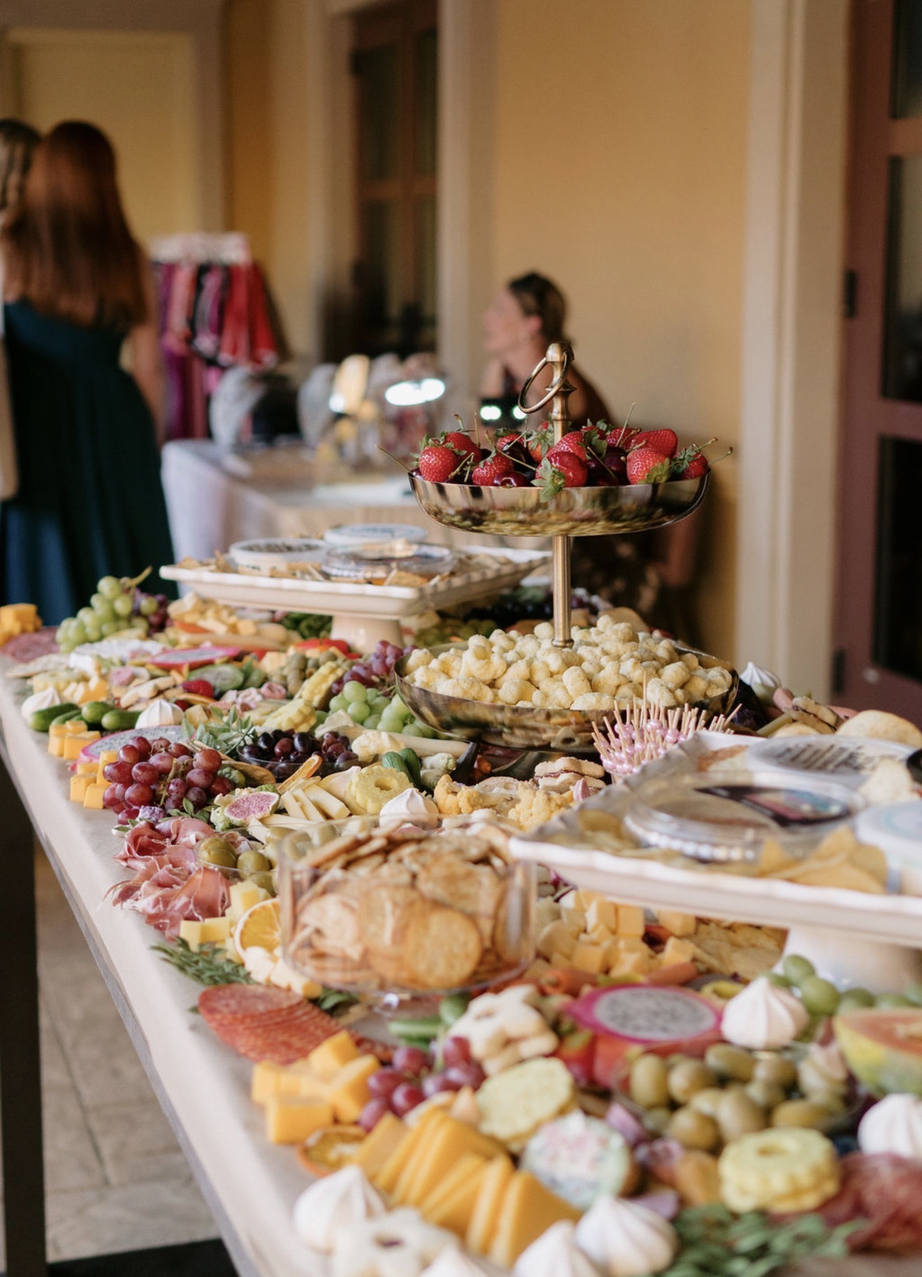 A table with various cheeses, fruits, and snacks, with people in the background in a warmly lit room.