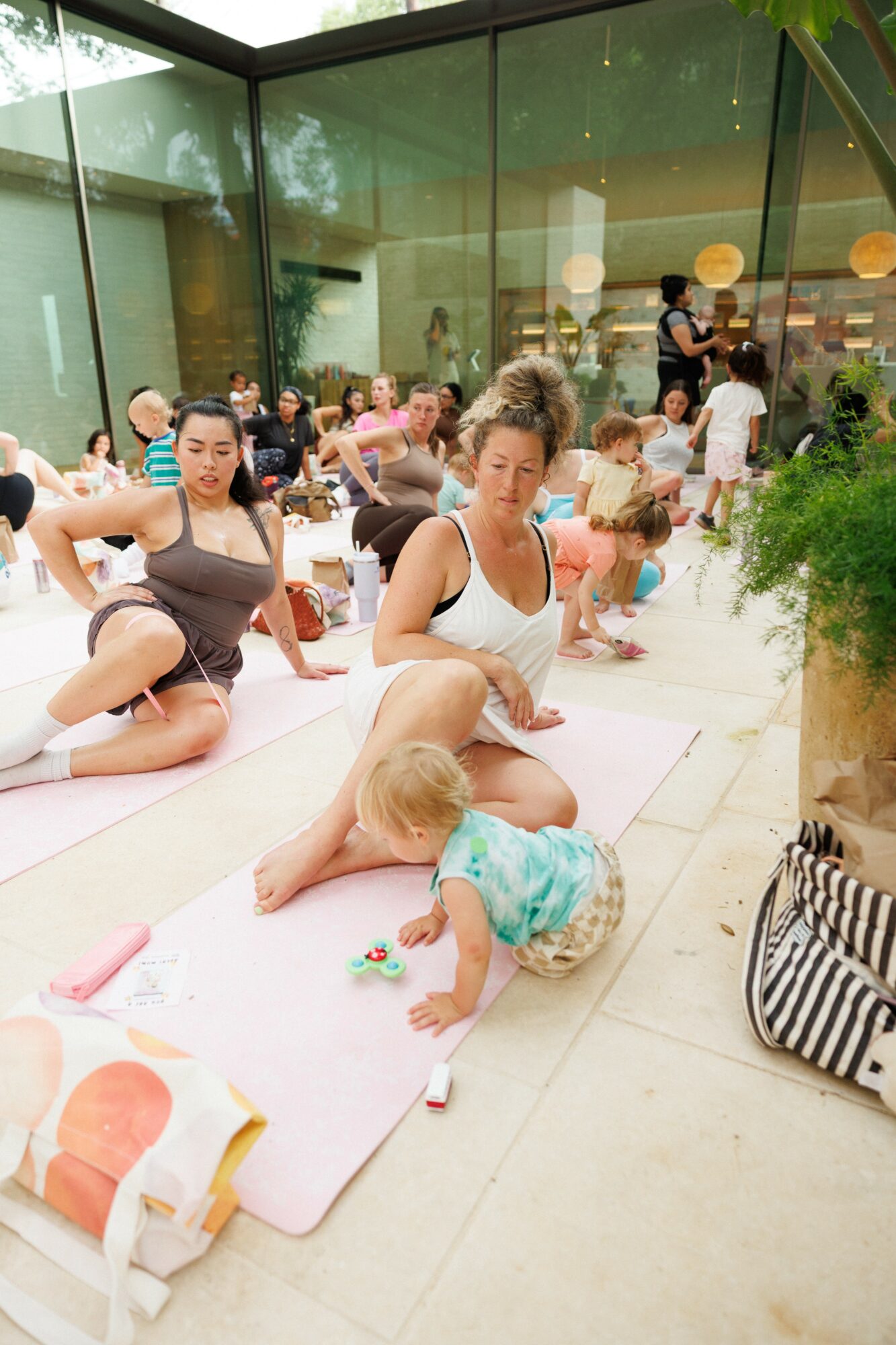 People sitting on mats indoors, children playing, and a large potted plant nearby.
