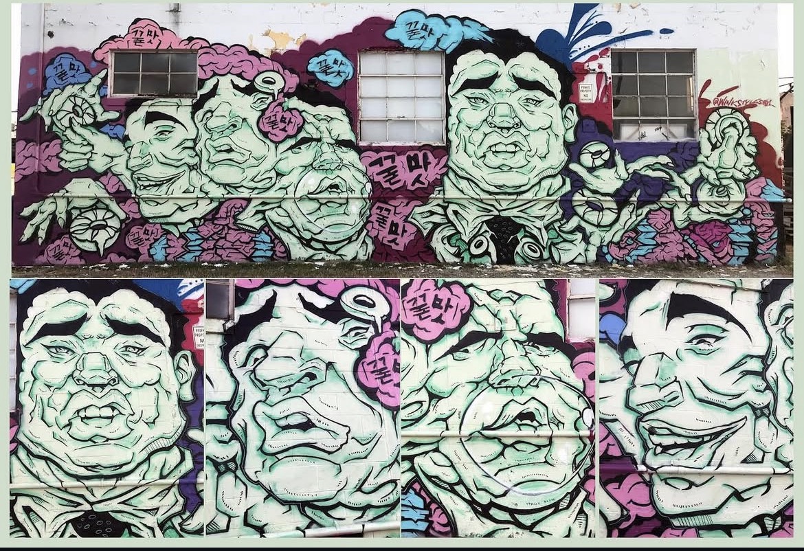 Colorful graffiti mural with large faces, clouds, and abstract shapes on a wall, featuring vibrant pink, blue, and purple colors.