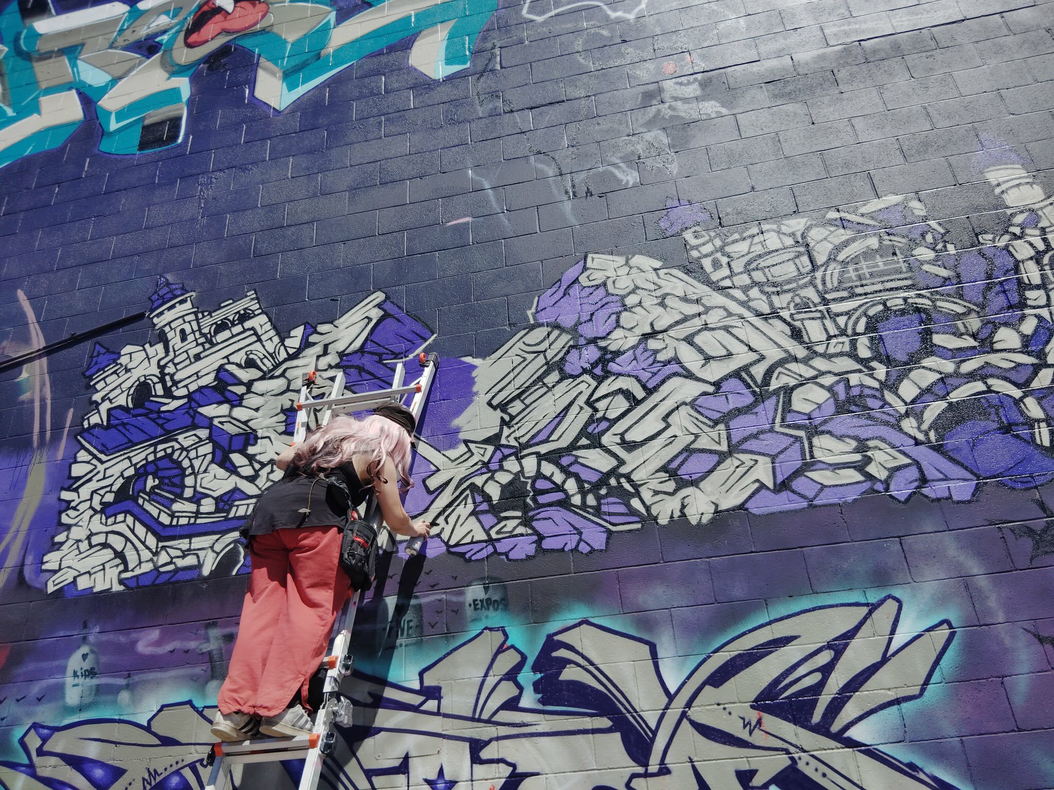 Person painting graffiti on a brick wall with colorful designs and lettering, wearing a backpack and red pants.