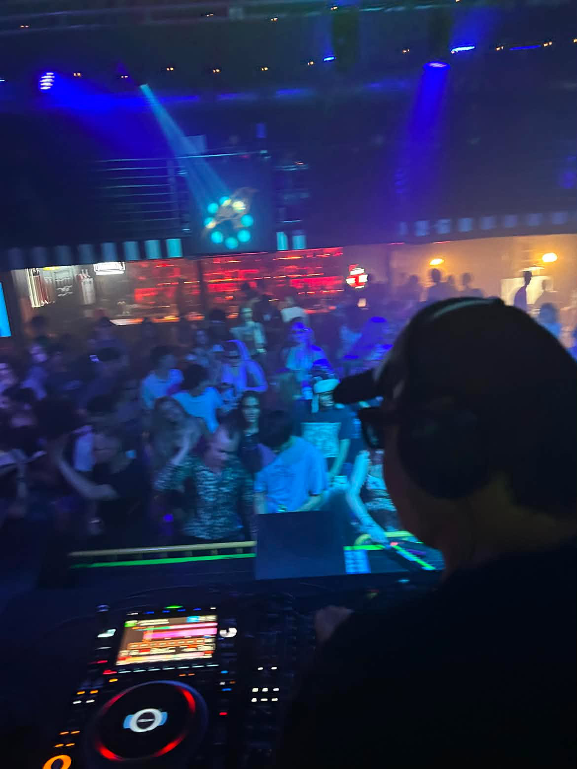 DJ wearing headphones in front of a crowd at a nightclub with colorful lights and a dance floor.