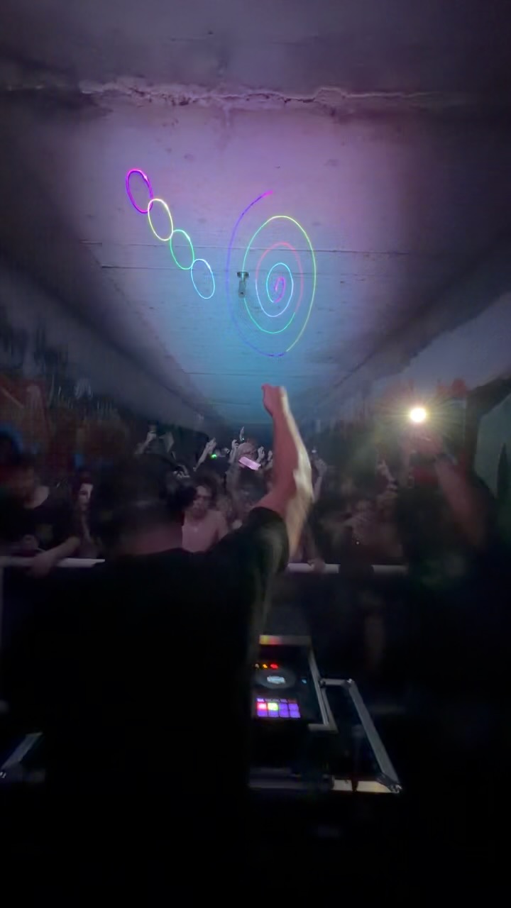 Crowd at a concert with a DJ, colorful light projections on the ceiling, and a person raising a hand.