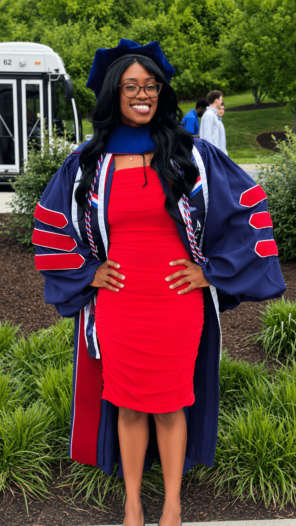 Young woman in graduation gown and cap, smiling outdoors with trees and a bus in background.