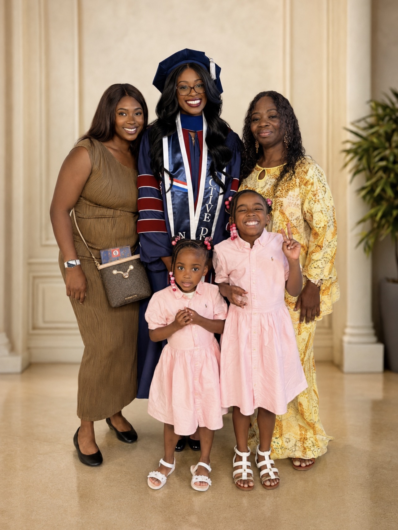 Group of six women and children in formal attire, standing indoors with a cream-colored wall and plant in background.