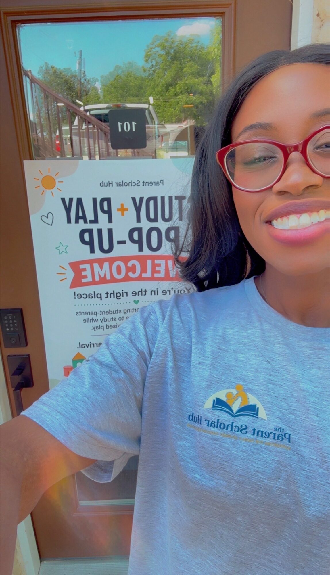 Young woman with glasses smiling, wearing a blue shirt, standing indoors near a window with a sign behind her.